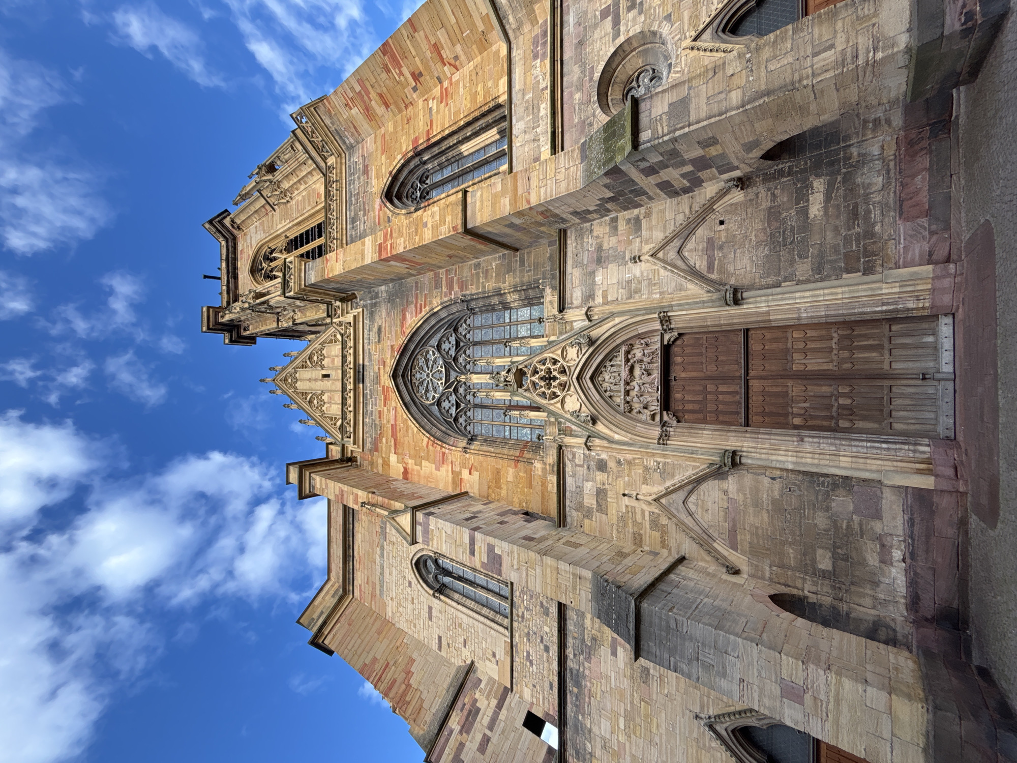 The image shows a low-angle, wide shot of the Saint Martin's Church in Colmar, France, on a bright, partly cloudy day. The church is constructed of sandstone with red and yellow hues and features Gothic architectural elements, including pointed arches, ornate windows, and a tall bell tower. The main entrance has large wooden double doors beneath a carved tympanum and a rose window. The sky above is a vibrant blue with scattered white clouds. The ground in front of the church is paved with cobblestones. There are no people visible in the image.