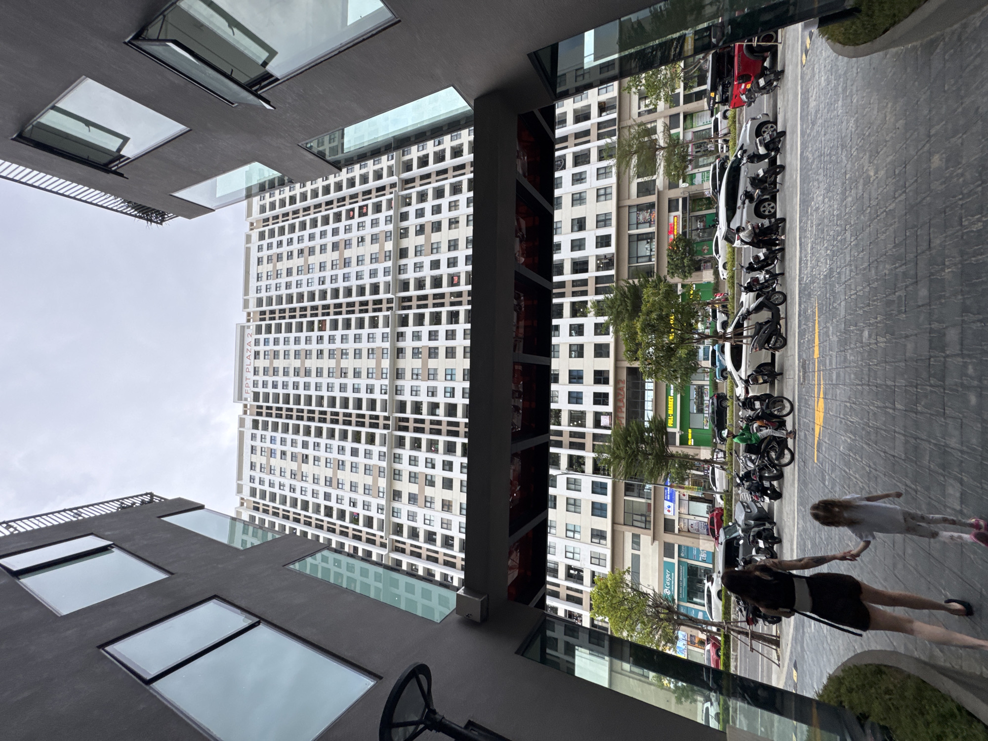 In Đà Nẵng, Vietnam, a wide-angle shot captures the perspective from a ground-level courtyard looking up at a tall, modern apartment building. The foreground features two people, a woman and a child, walking away from the camera towards the street. The woman, wearing a black dress and heels, holds the child's hand, who is dressed in a white t-shirt and patterned leggings. A line of parked motorcycles and scooters is visible along the curb, with a few cars further down the street. The mid-ground reveals the lower floors of commercial buildings with storefronts and trees lining the street. The background is dominated by the immense apartment complex, characterized by its numerous balconies and windows, under an overcast sky. The architecture suggests a contemporary urban environment.