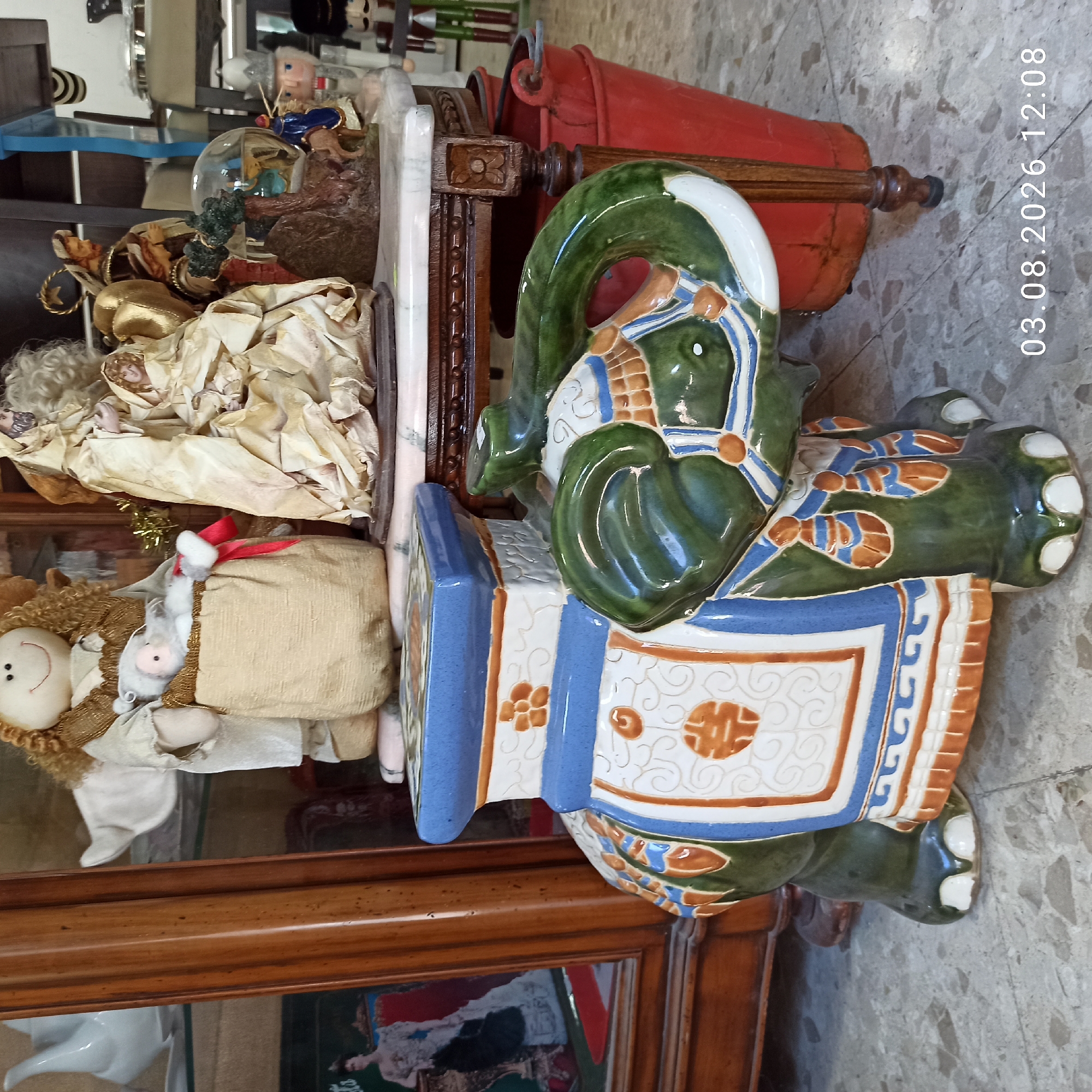 A ceramic elephant figurine, painted in green, blue, and orange, is positioned in the foreground of an indoor setting. The elephant has a flat, rectangular top, suggesting it might function as a small table or stool. Behind the elephant, a wooden cabinet with glass doors is visible, containing various decorative items including a nativity scene with figures of Mary, Joseph, and a baby, and an angel figure holding a baby. To the right of the cabinet is a wooden table with a marble top, on which rests a glass snow globe containing a nativity scene. Adjacent to the table, two red buckets are stacked. In the background, nutcracker figures stand on a shelf. The flooring appears to be tiled. The image was captured on August 3, 2026, at 12:08 PM. This scene is located in Ruvo di Puglia, Italy.