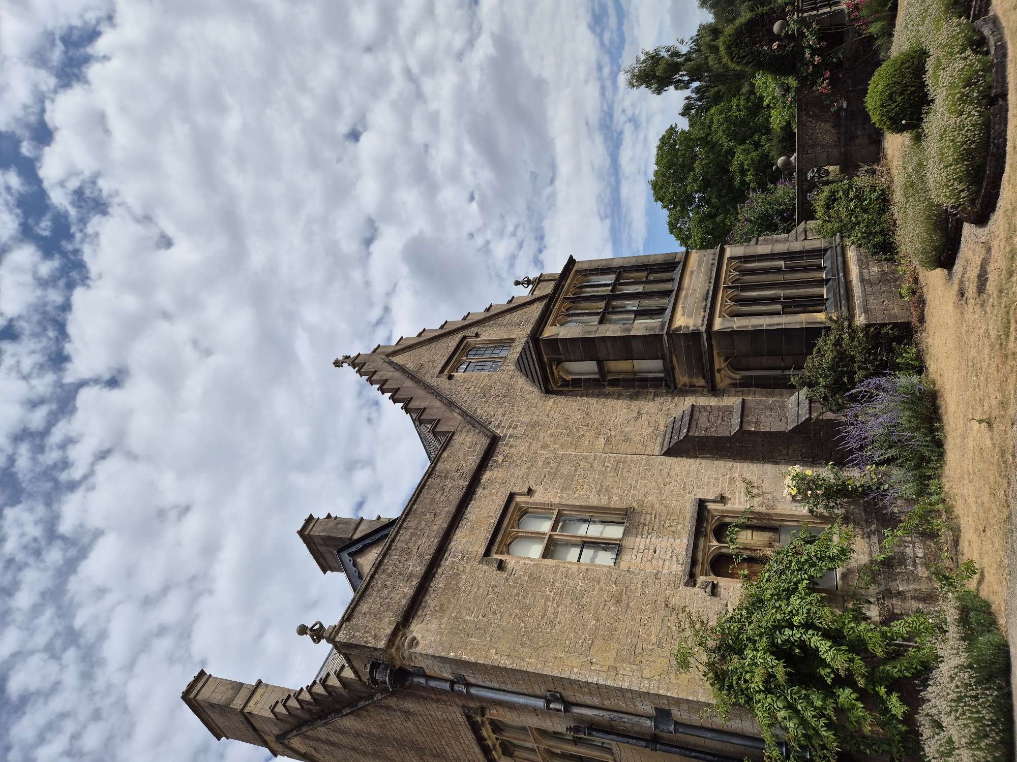 This is an exterior, low-angle, full shot of a large, stone building, likely a historic house, located in the city of Nottingham, United Kingdom. The building is constructed from yellowish-brown stone blocks and features several Gothic-style architectural elements, including pointed arch windows with leaded panes, crenellations along the roofline, and a prominent bay window. Two tall chimneys rise from the roof. Verdant greenery, including climbing vines and various flowering plants such as lavender, are visible in the foreground and around the base of the building. A dry, light-brown lawn covers the ground in front of the house. In the background, lush green trees and a stone wall are present. The sky above is partly cloudy, with large, white cumulus clouds against a blue sky. There are no people or animals visible in the image. The scene appears to be an estate or a historical landmark on a bright, possibly sunny day.