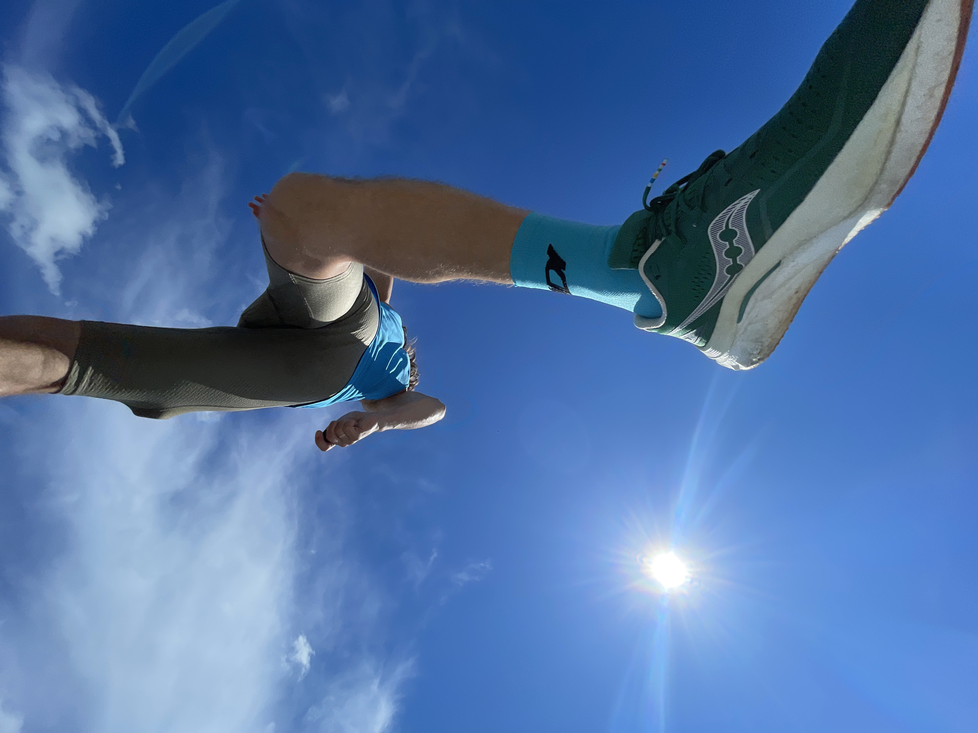 An outdoor, low-angle, extreme close-up shot captures a person's legs and feet against a bright blue sky with scattered clouds and a visible sun with lens flare. The individual is wearing athletic compression leggings, blue athletic shorts, bright teal socks with a black logo, and dark green running shoes with a white sole. One shoe is in the foreground, while the other leg is positioned higher up and to the left. The person's hand is visible near their torso on the left side of the frame, with their fist closed. The sun, located in the lower left quadrant of the image, is intensely bright and appears to be low in the sky, possibly suggesting a sunrise or sunset. The overall impression is one of dynamic movement or mid-action, with the focus on the lower body against a vast sky.