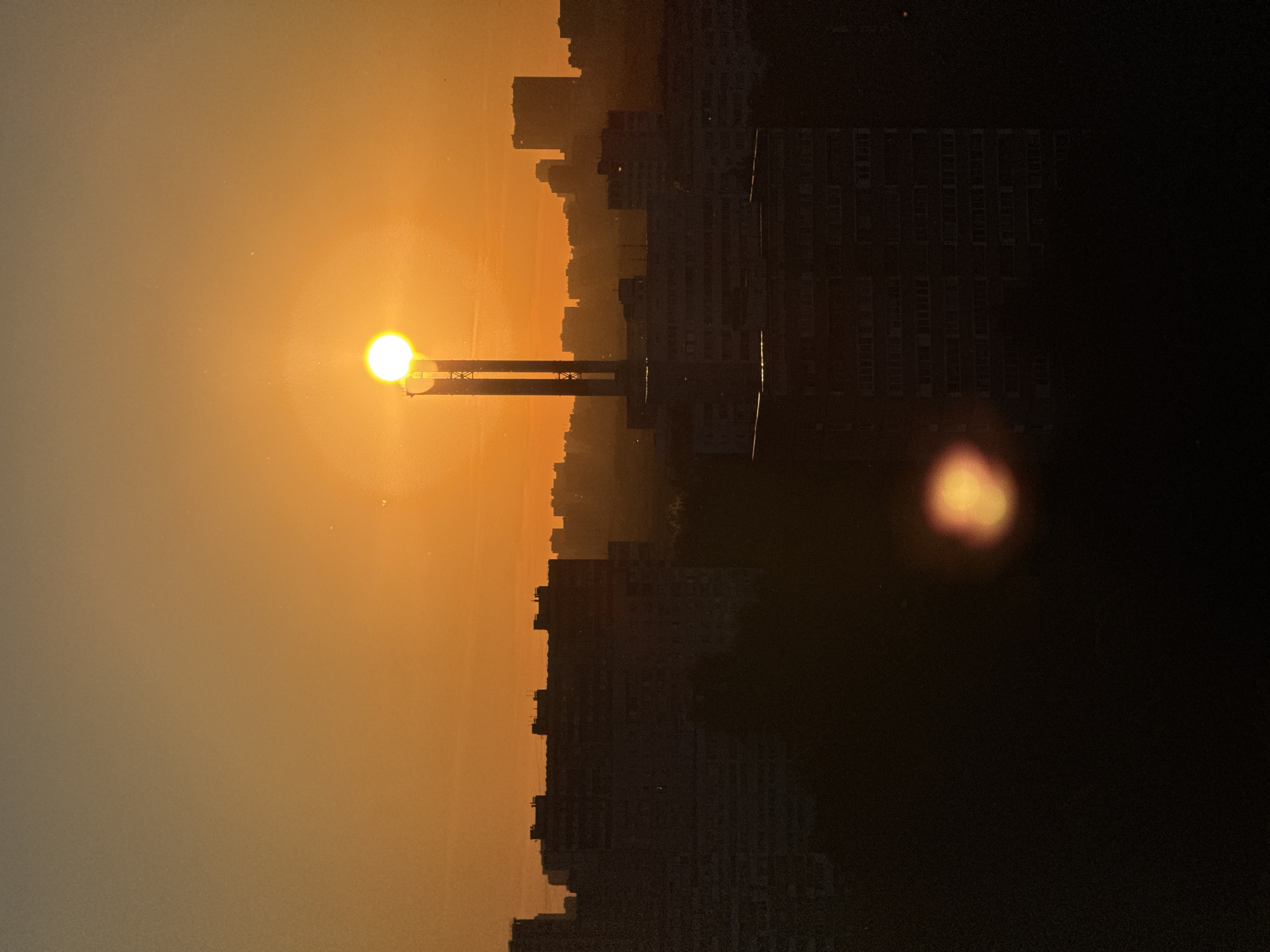 The image captures an urban landscape at either sunrise or sunset, characterized by an intensely bright, circular sun positioned between two tall, vertical industrial structures. The sky exhibits a warm gradient, transitioning from a vivid orange around the sun to a lighter, somewhat hazy orange-brown higher in the frame. The entire lower two-thirds of the image, encompassing the cityscape, is rendered in deep silhouette due to the strong backlighting.
The foreground and mid-ground are dominated by the dark silhouettes of numerous multi-story residential buildings, likely panel-style apartment blocks, which form a dense, jagged horizon line against the illuminated sky. Additional, less defined building silhouettes extend into the hazy background, suggesting a vast urban sprawl. No individual people or explicit actions are discernible. A prominent, diffuse orange-yellow lens flare is visible in the lower central-right portion of the image. The architectural style of the high-rise residential buildings and the industrial chimneys are characteristic of urban infrastructure found in Moscow, Russia. Small, scattered specks, consistent with dust or sensor artifacts, are observable against the sky.