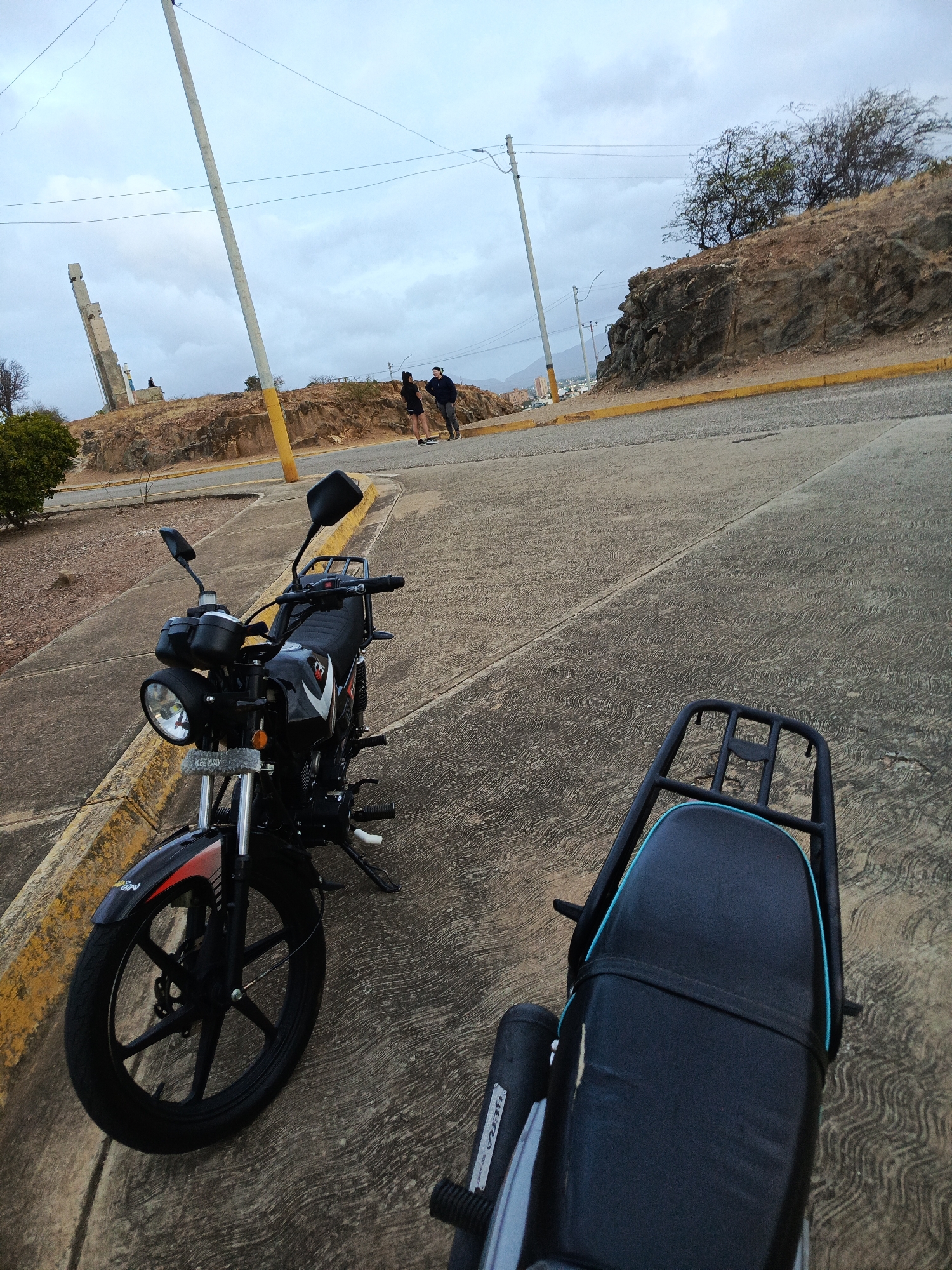 The image depicts a street scene in Juan Griego, Venezuela, featuring two motorcycles parked on a concrete road. The first motorcycle, a black Keeway, is partially visible in the foreground. The second motorcycle, with a black seat trimmed in turquoise, is positioned closer to the viewer. In the background, two individuals are walking along the road. A hillside with sparse vegetation and a yellow curb runs parallel to the road. A concrete monument stands on the left side of the image against a cloudy sky. Utility poles are visible in the distance.