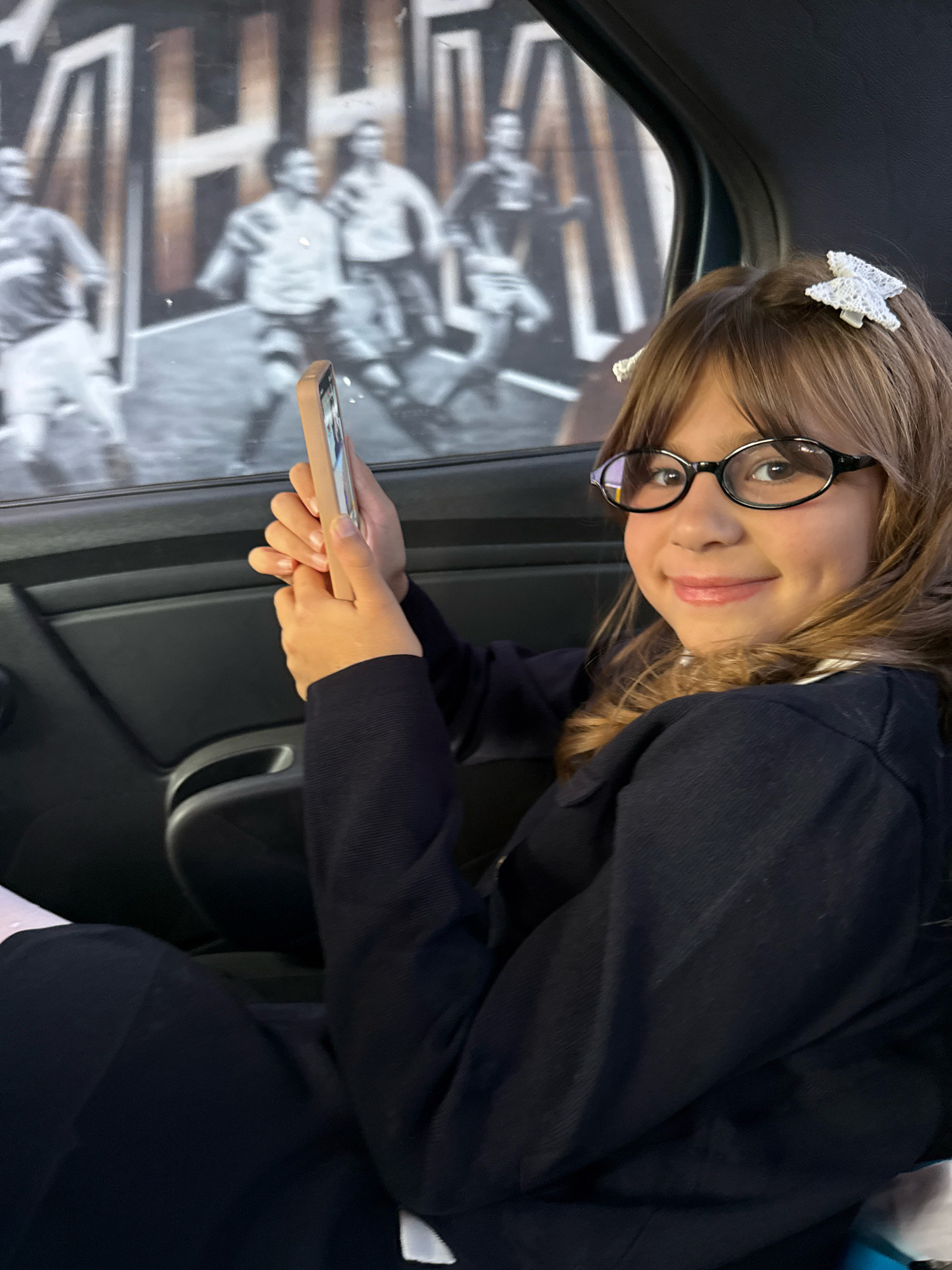 A young girl with blonde hair, wearing glasses and a navy blue long-sleeved shirt, is seated in the back of a vehicle. She is holding a mobile phone in her hands and appears to be looking at it or taking a picture. The background visible through the car window is a black and white mural depicting several figures in motion, possibly athletes. The interior of the car, including the door panel, is dark gray. The scene suggests a casual moment captured inside a moving vehicle. There is no information in the image to suggest the location is Rusanovo, Russia.