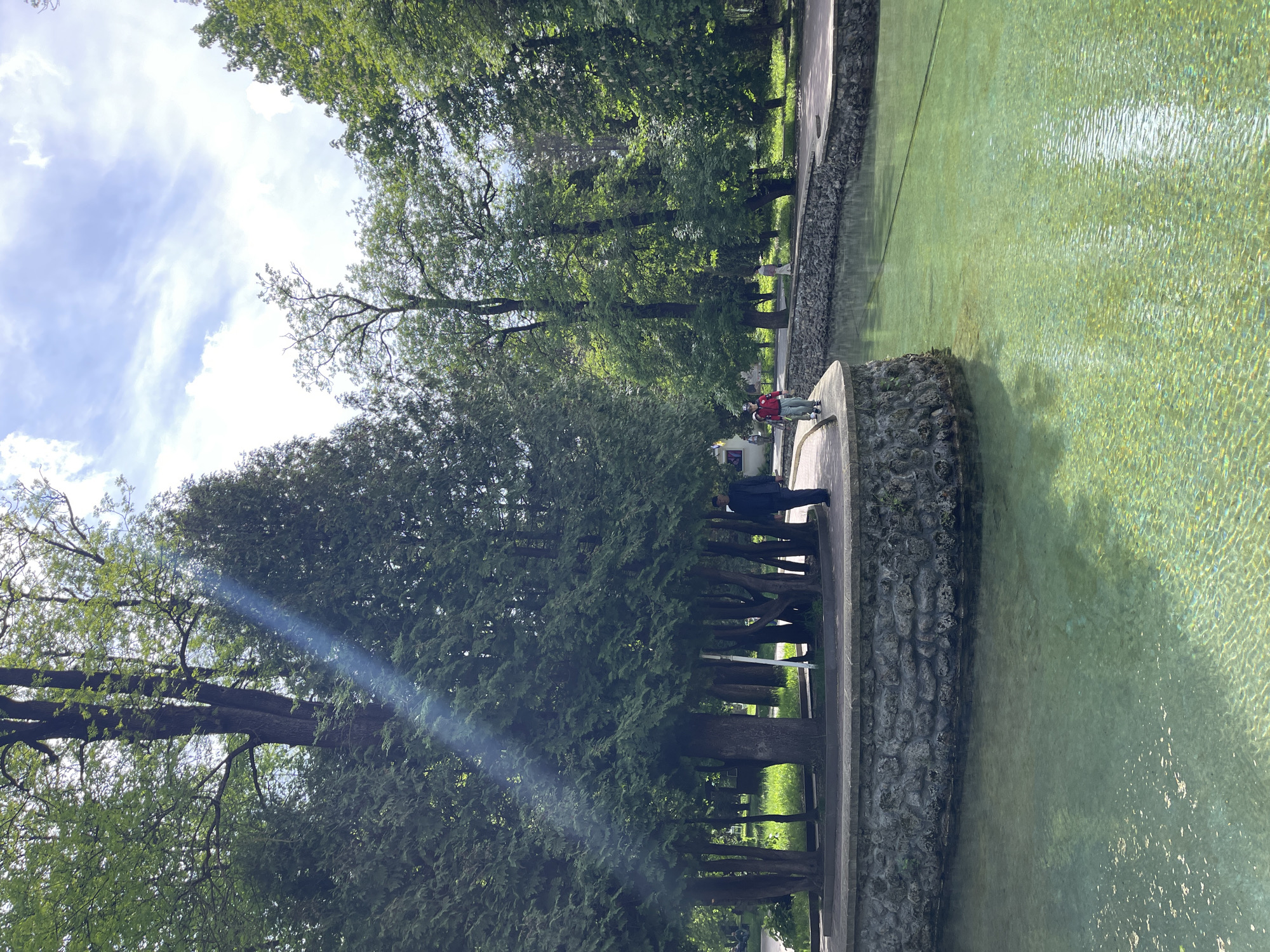 The image depicts a park in Kislovodsk, Russia. A clear, shallow water body is visible on the right. A stone wall forms the border. Along the wall on the left is a paved walkway with trees growing beside it. Several people are visible on the walkway. The sky above is partially cloudy with visible sunlight.