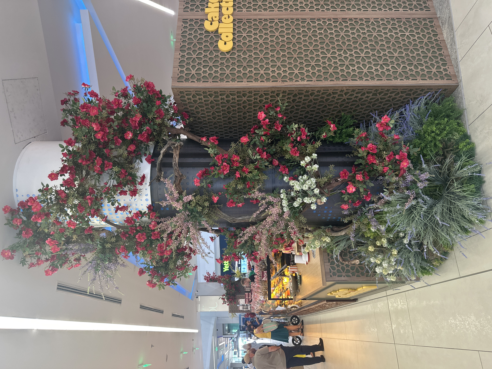This image captures a detailed view of a retail environment within Paradise Center, Sofia, Bulgaria. The central focus is a kiosk or cafe stall wrapped around a large, circular structural pillar, which is elaborately decorated with extensive artificial flora.
The pillar is adorned with numerous clusters of vibrant red roses, delicate pink and purple flowering branches resembling cherry blossoms and lavender, and small white flowers, all intertwined with faux dark wood branches and dense green foliage. Portions of the pillar also display a dark, perforated grid-like surface. At the base of this decorative pillar, a ground-level arrangement of artificial green plants, lavender, and other small white flowers creates a lush, garden-like foundation.
To the right, a tall, decorative screen serves as a partial wall for the kiosk. This screen is constructed from light brown wood featuring an intricate, repeated geometric cutout pattern, with a dark green backing visible through the cutouts. Mounted in yellow, three-dimensional lettering on this screen is the brand name "Cake Collection."
The kiosk features a light-colored wooden counter displaying various cakes, pastries, and sweets in glass cases. Small white cups are stacked on the counter. Behind the counter, a male employee in a dark shirt is visible. On the left side of the counter, several customers are present. An older man in a light brown short-sleeved shirt and dark trousers stands with his back mostly to the camera, facing the display. A woman in a yellow top and dark skirt stands near a small, wheeled cart, looking at the items. Another individual is partially obscured near the counter.
The floor consists of large, reflective light-colored square tiles. The white ceiling is equipped with linear strip lighting and sections of blue accent lighting. Visible on the ceiling are multiple air conditioning vents. In the background, beyond the kiosk, other retail spaces are discernible, including a store with a prominent green sign displaying the word "Yett."