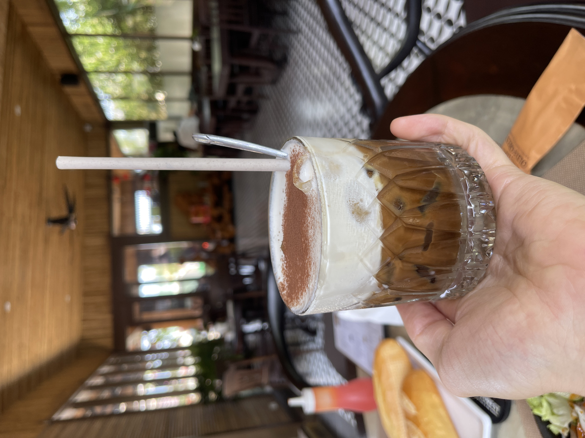 A close-up, rotated image shows a person's left hand holding a clear, textured glass of a layered iced drink, likely coffee, within a cafe setting. The drink features a dark brown, icy base topped with a thick, foamy white layer, generously dusted with cocoa powder on one side. A light-colored straw and a small metallic spoon are inserted into the drink.
The background reveals a brightly lit cafe interior during the daytime. To the left, wooden paneling forms a wall, while large windows extend across the upper section, offering a blurred view of lush green foliage and bright sunlight, creating a bokeh effect. A patterned floor or wall section is visible on the right. On the table, partially visible near the glass, are a golden-brown baguette, a small red sauce bottle, and some salad greens, suggesting a meal is being enjoyed. A piece of paper or cardboard, partially obscured, has the text "OLD COFFEE" visible on it. The overall atmosphere is relaxed and casual, characteristic of a daytime cafe visit in a tropical location like Phú Quốc, Vietnam.