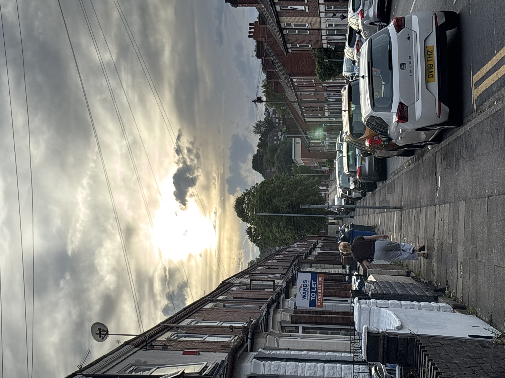 A street scene in Stoke-on-Trent, United Kingdom, shows a row of terraced houses lining a paved sidewalk. Parked cars are visible along the right side of the street, with license plates visible. An adult female walks along the sidewalk, facing towards the distant end of the street. A 'To Let' sign from a property management company (WARDS) is affixed to one of the houses. Overhead, power lines stretch across the cloudy sky, where the sun is obscured by cloud cover.