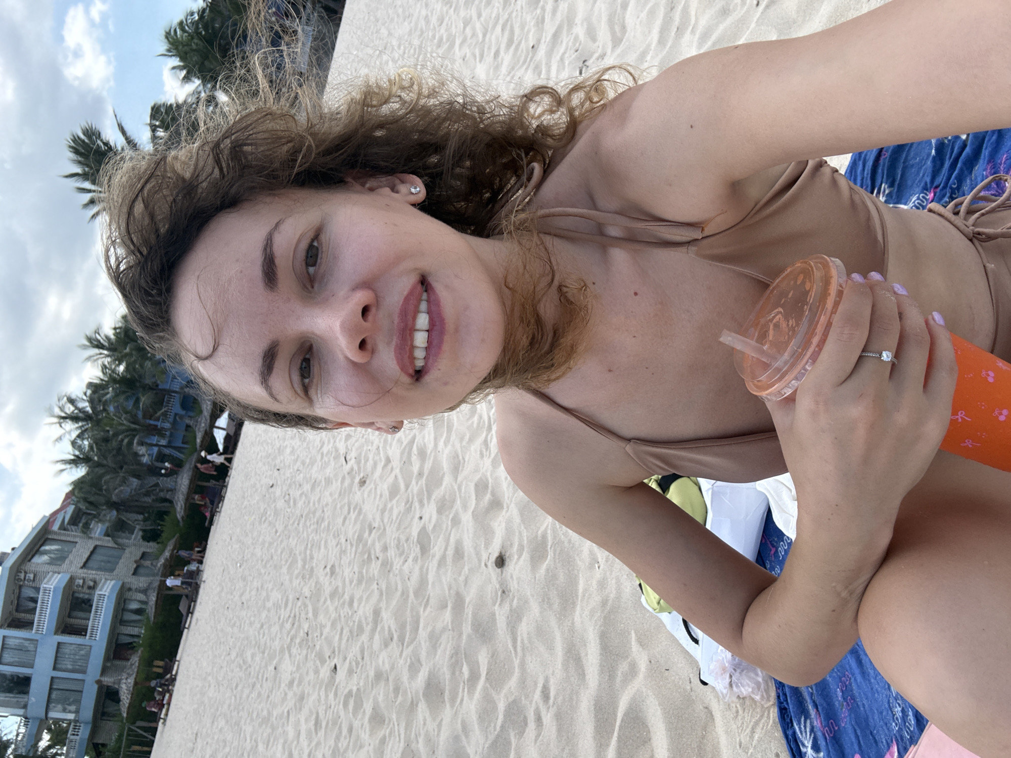 A woman with curly brown hair is sitting on a sandy beach in Mũi Né, Vietnam, holding an orange drink with a straw. She is wearing a beige bikini and a silver ring on her left hand. Behind her, the beach extends to the ocean, with palm trees and a light blue building in the background. Several lounge chairs and umbrellas are visible on the sand. The sky is partly cloudy.