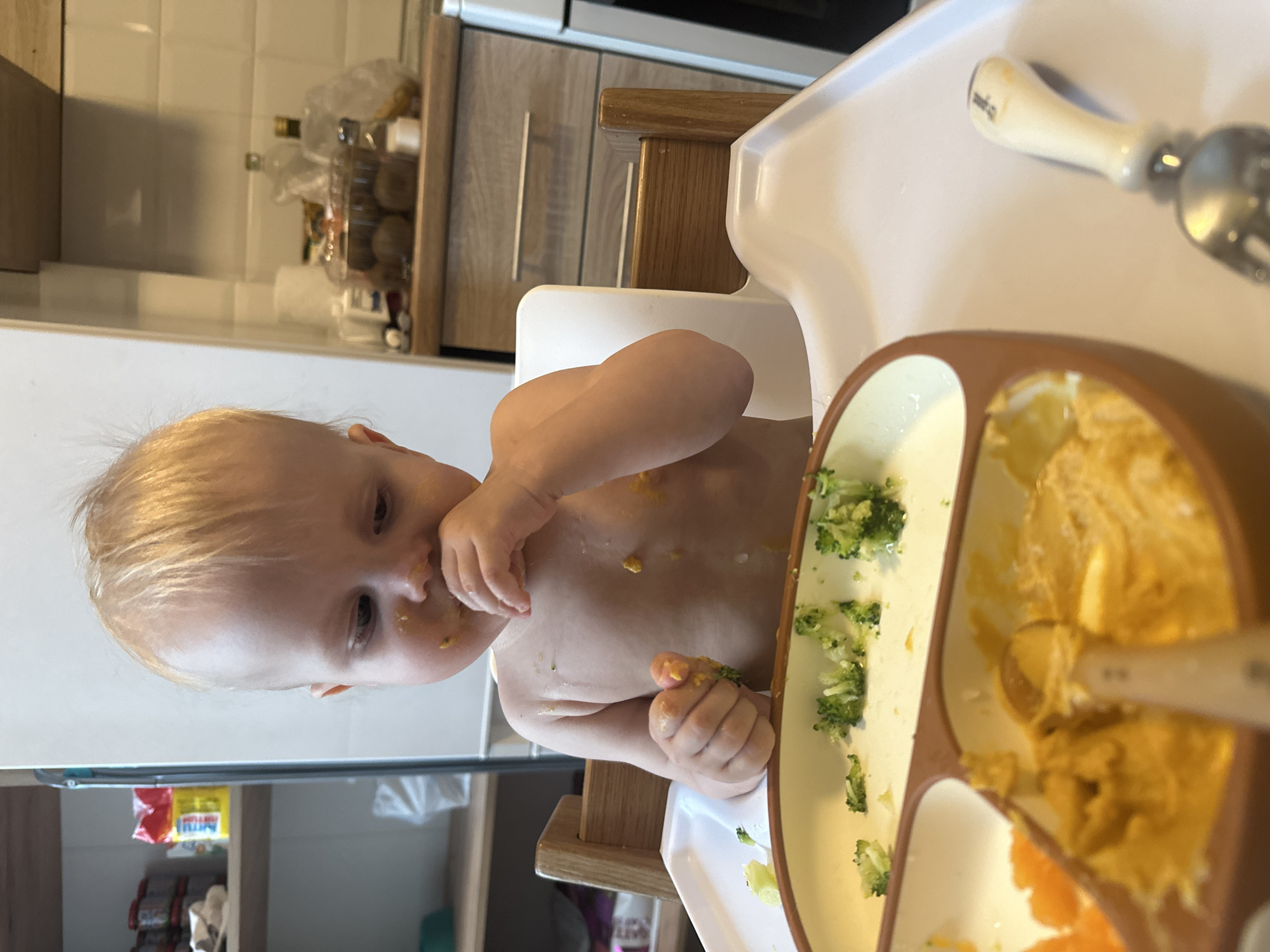 A young, fair-skinned child with blond hair is sitting in a high chair, eating. The child is partially undressed, with food smudged on their face and chest. In front of the child is a segmented plate containing mashed yellow food (likely squash or sweet potato), pieces of broccoli, and orange segments. A spoon is visible in the plate. The high chair is white with a wooden frame. The background shows a kitchen area with light-colored cabinets and some items on shelves. The location in the city of Khimki, Russia is not discernible from the image.