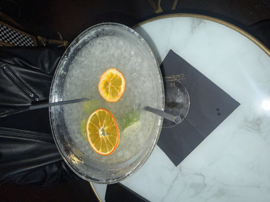 A large, wide-rimmed glass cocktail, filled with crushed ice, a pale liquid, multiple lime wedges, and two dried orange slices of differing diameters, is centrally positioned. Two dark straws, one vertical and one angled, are inserted into the drink. The glass is partially held by an individual whose torso, clad in a black leather jacket, is visible in the upper background. The cocktail rests on a black square napkin, imprinted with the white text "LE CAROUSEL" vertically along its right edge. This napkin is on a round table with a white marble top and a gold-colored rim. The background is dark and out of focus, revealing a faint black and white checkered pattern in the upper right. The "LE CAROUSEL" branding indicates the location as the brasserie Le Carousel, situated at 232 Rue de Rivoli, 75001 Paris, France.