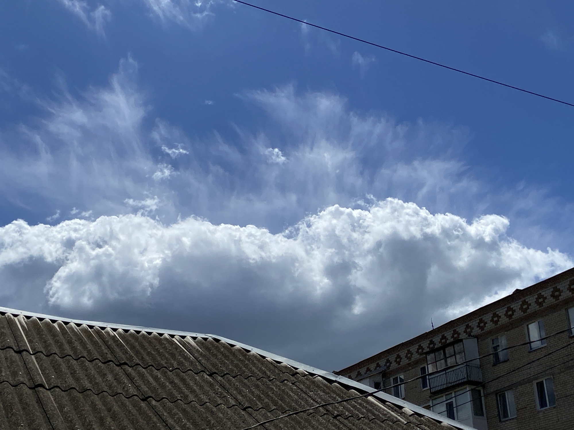 The image shows a sky with cirrus and cumulus clouds above two buildings in an urban environment. The sky is primarily blue with wispy cirrus clouds stretching across the upper portion. A band of large, puffy cumulus clouds extends horizontally across the middle of the image. In the foreground, on the lower left, is the gray, corrugated roof of a building. To the right is a brick apartment building with visible windows and a balcony. A power line stretches across the upper right corner of the frame. The location is Unknown, Unknown. There are no visible people.