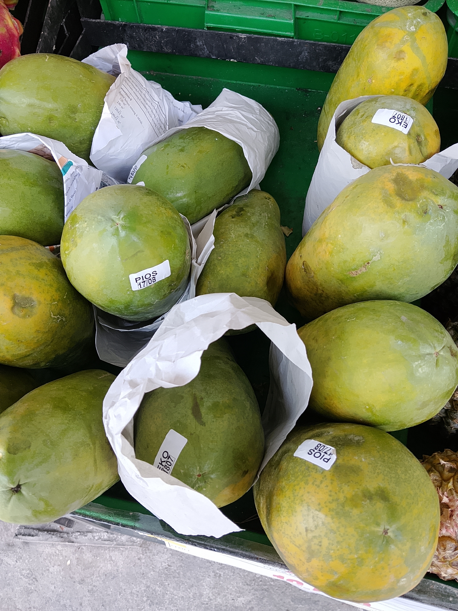 A close-up, overhead shot displays a pile of green and yellow papayas, some wrapped in white paper, in a green plastic crate. The papayas vary in ripeness and size. Several have white stickers with black text, one reading "PIOS 17/08" and another "EKO 1807". The crate is situated on a textured, grey surface, possibly a market stall. The background shows more green crates. This scene is located in Karangboyo, Indonesia.