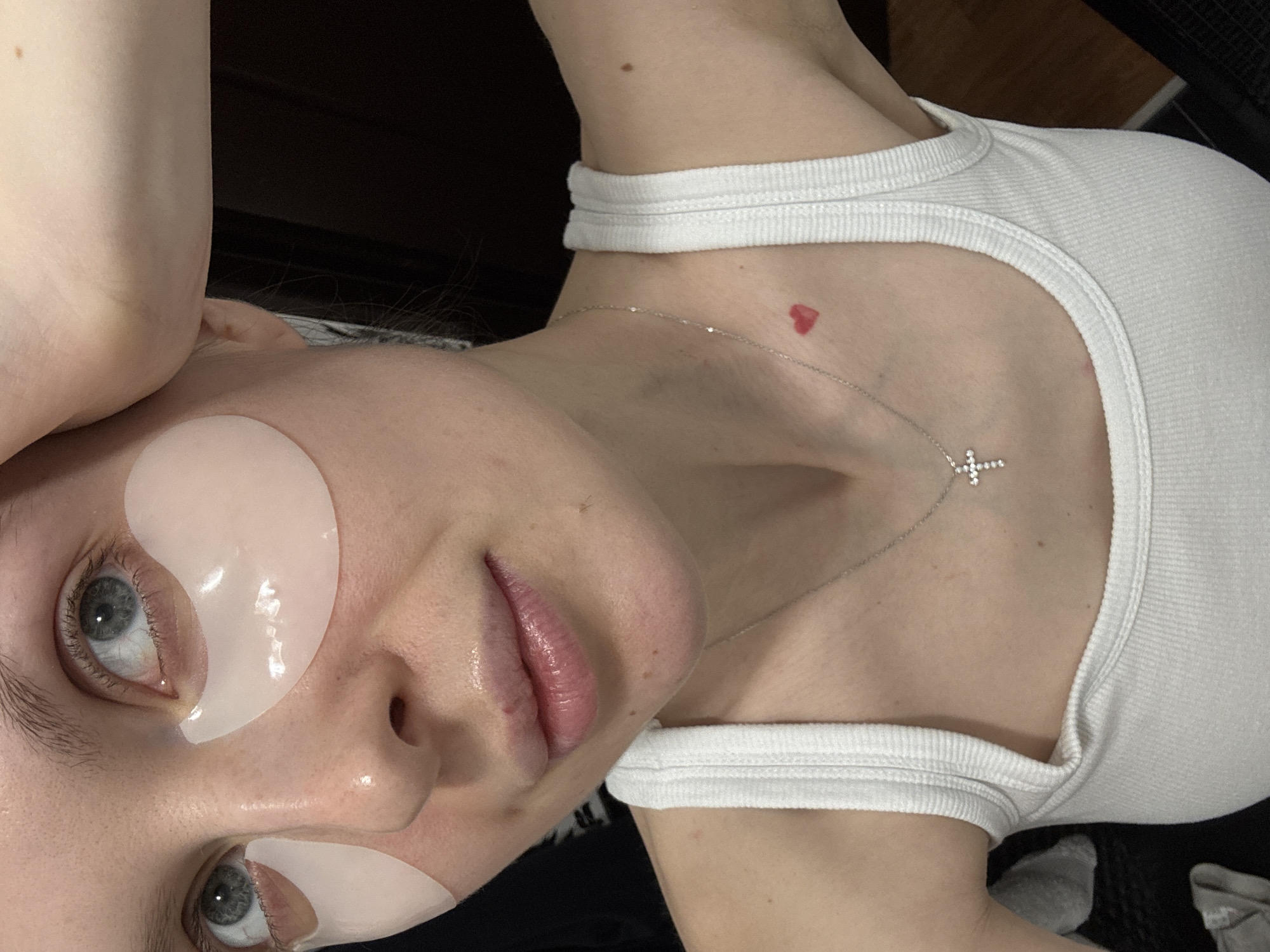 A young person is positioned at an angle, wearing eye masks under both eyes, and a white tank top. A silver chain necklace with a cross pendant is visible around their neck. A red heart-shaped mark is present on their chest. The background is dark, possibly indicating an indoor setting. There is no indication of Moscow, Russia.