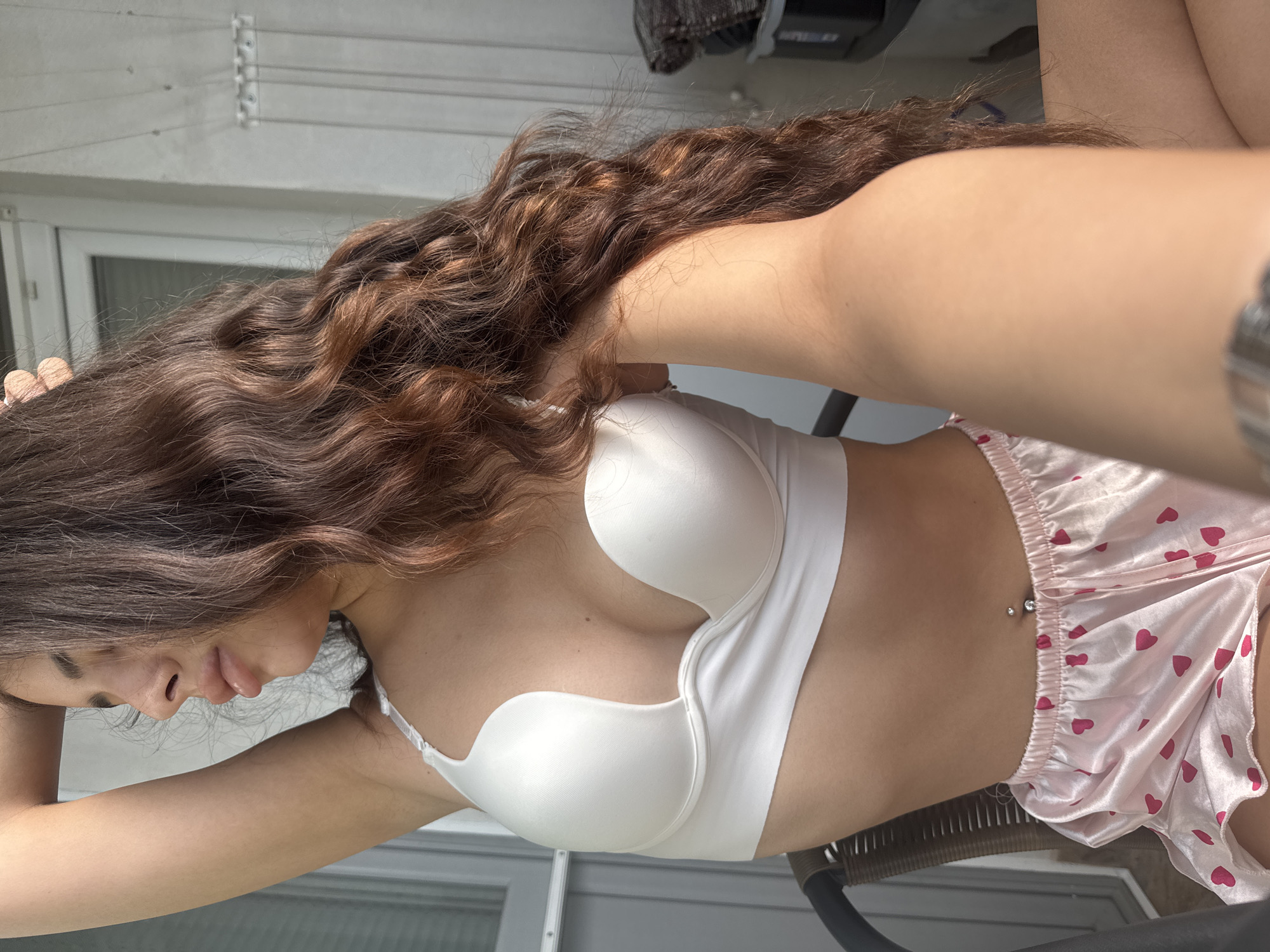 A young woman with long, wavy brown hair is seated outdoors on a chair, facing right and looking away from the camera. She is wearing a white strapless bra and pink satin shorts with a heart pattern. Her left arm is raised above her head, and her right arm is extended forward, holding a phone out of frame, presumably for a selfie. A silver belly ring is visible on her navel. The background includes a building with windows and exterior walls, suggesting an urban setting, possibly a balcony or patio. The lighting indicates it is daytime, with sunlight casting shadows. The provided location of Plovdiv, Bulgaria, is not discernible from the visual elements of the image itself.