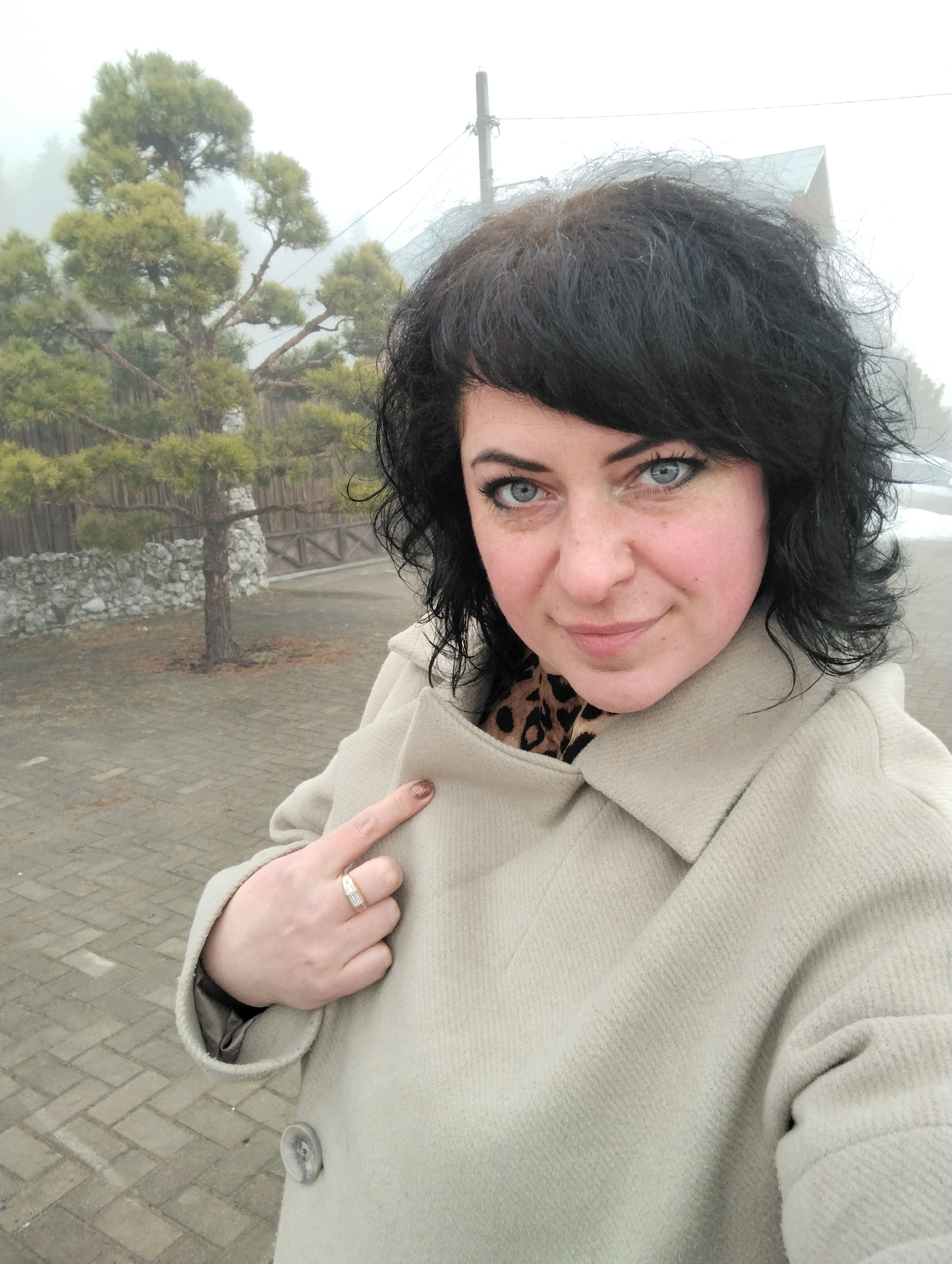 A selfie-style photograph shows a woman with blue eyes and dark curly hair, wearing a beige coat and a leopard print scarf. She is pointing at her coat with her right index finger, on which she wears a ring. The background features a tree covered in mist, a wooden fence, and a stone wall. The ground is paved with interlocking bricks. The photo appears to have been taken outdoors on a cloudy day. Although the exact location is not specified, the query mentions the city of Votkinsk, Russia, suggesting the photo was taken there. No specific actions or interactions are depicted apart from the woman pointing.