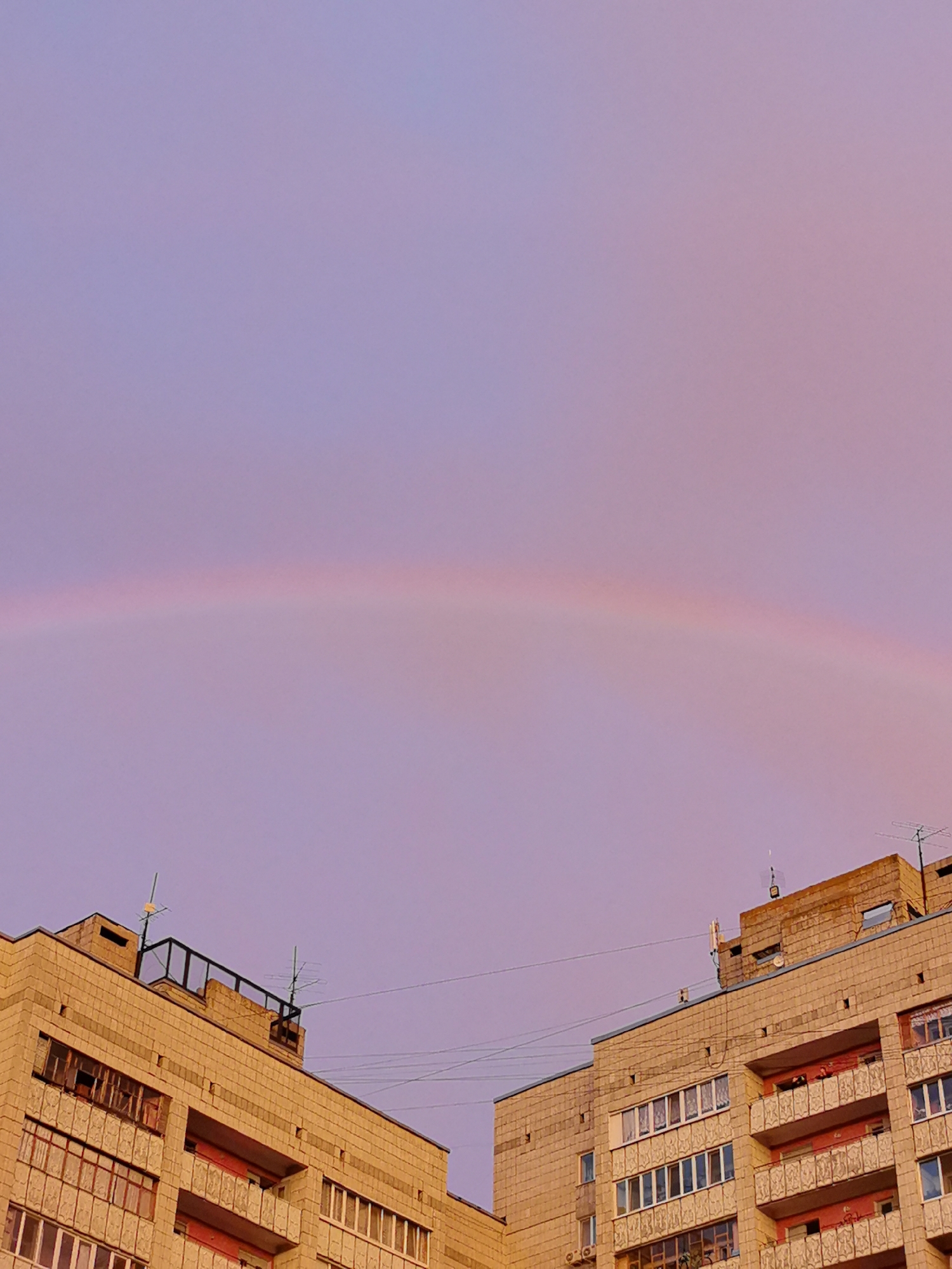 The image depicts a low-angle view of apartment buildings against a pastel purple sky with a faint rainbow arching across it. The buildings, constructed with beige brickwork, feature multiple floors with balconies and windows. Some windows are broken, and others appear to have curtains. Antennas and other rooftop fixtures are visible on the buildings. The scene appears to be set in a residential area of Kazan, Russia, likely during twilight or just after a rainfall. No people are visible in the frame.