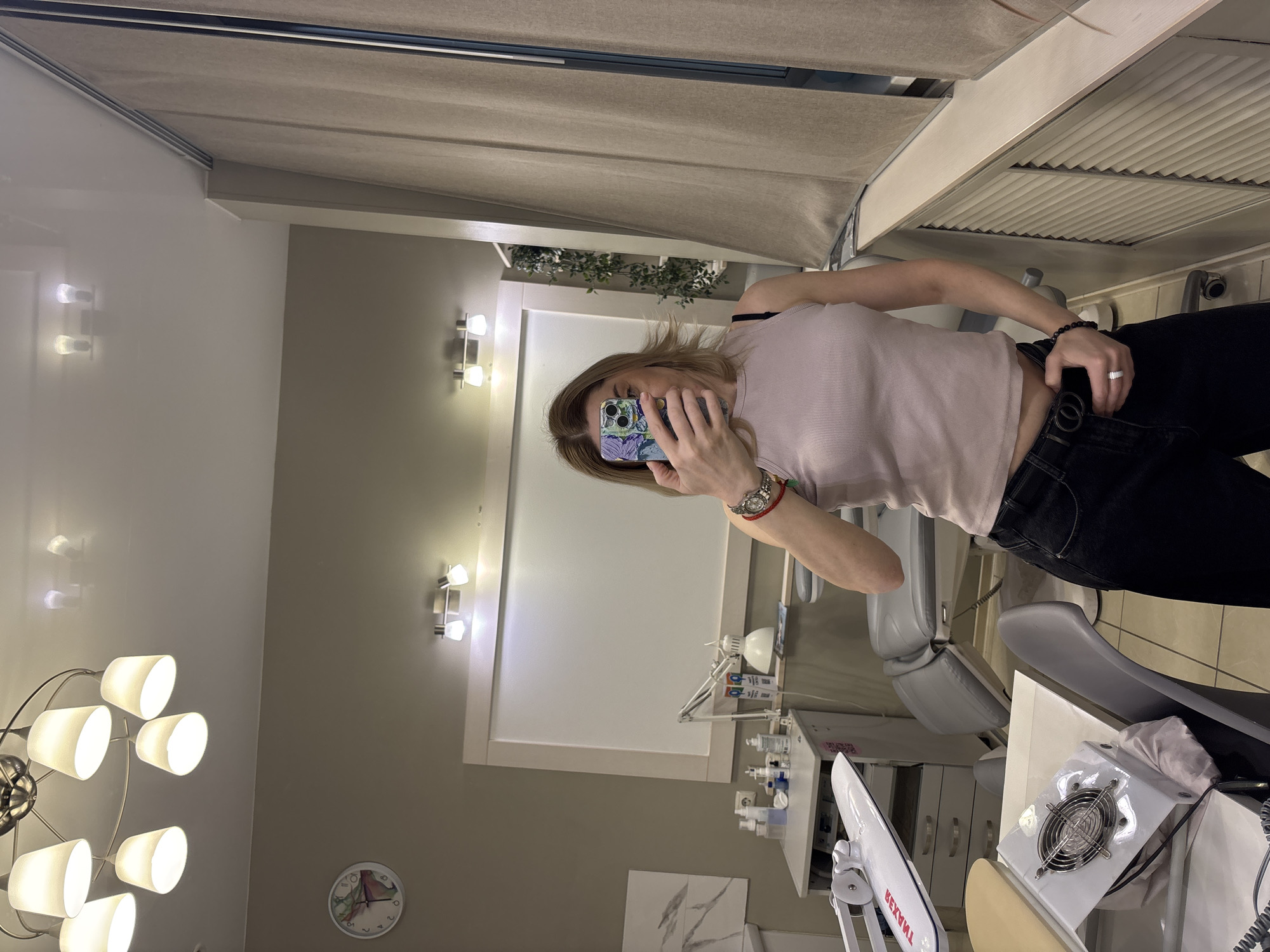 A woman is taking a selfie in what appears to be a medical or dental clinic. The woman is wearing a light pink top and black pants. She is holding a phone with a patterned case. In the background, there is a dental chair, medical equipment, wall-mounted lights, and a clock. The location is presumed to be in Vsevolozhsk, Russia.