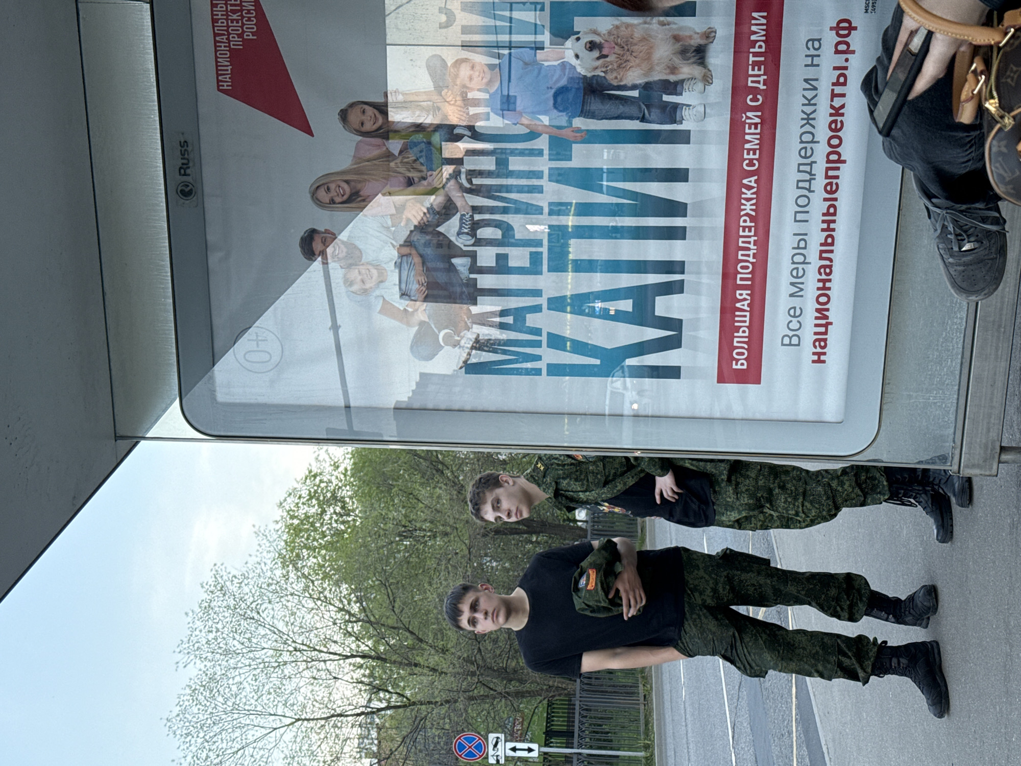 The image depicts two young men dressed in military fatigues standing on a sidewalk next to a bus stop in what appears to be Moscow, Russia. The bus stop has a large advertisement showcasing a family with children and a dog, promoting state support for families with children, with the text "МАТЕРИНСКИЙ КАПИТАЛ" and a website address. One of the men is holding a military cap. The background shows trees with fresh green leaves, suggesting it is spring. A "No Parking" road sign is also visible.