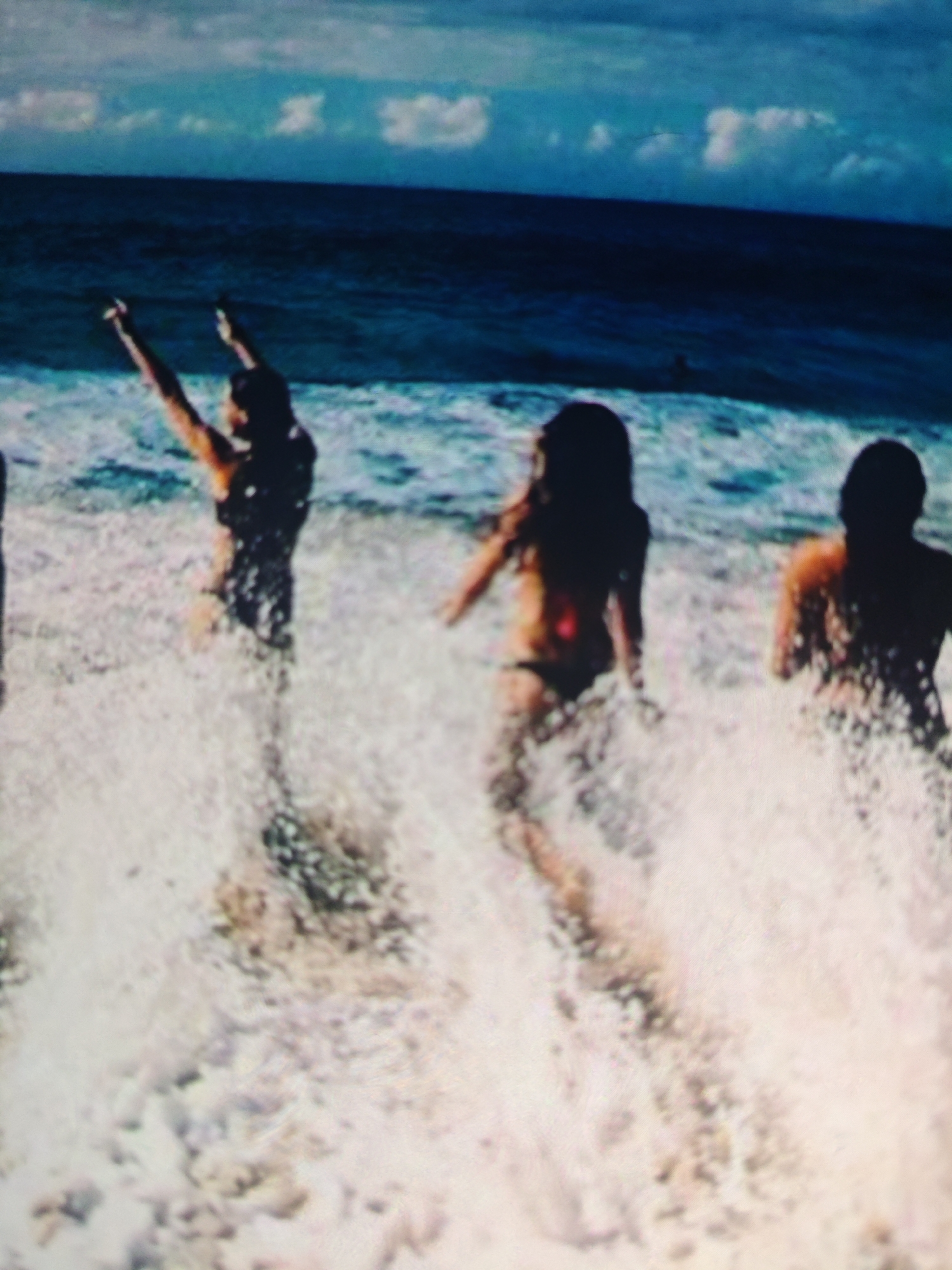 The image depicts a group of people in the ocean, with waves and spray visible around them. The sky is partly cloudy, with a mix of blue and white. The people are silhouetted against the bright water and sky, making it difficult to discern their features. One person in the foreground on the left appears to be raising their arms. To the right, two figures are more distinctly visible, one in a bikini, and another silhouetted person further to the right. The scene suggests a recreational activity in the water. The image quality is low, with a grainy texture and blurred details. There is no information within the image or the provided crops that would indicate a location in Krasnaya Polyana, Russia, or any urban setting.