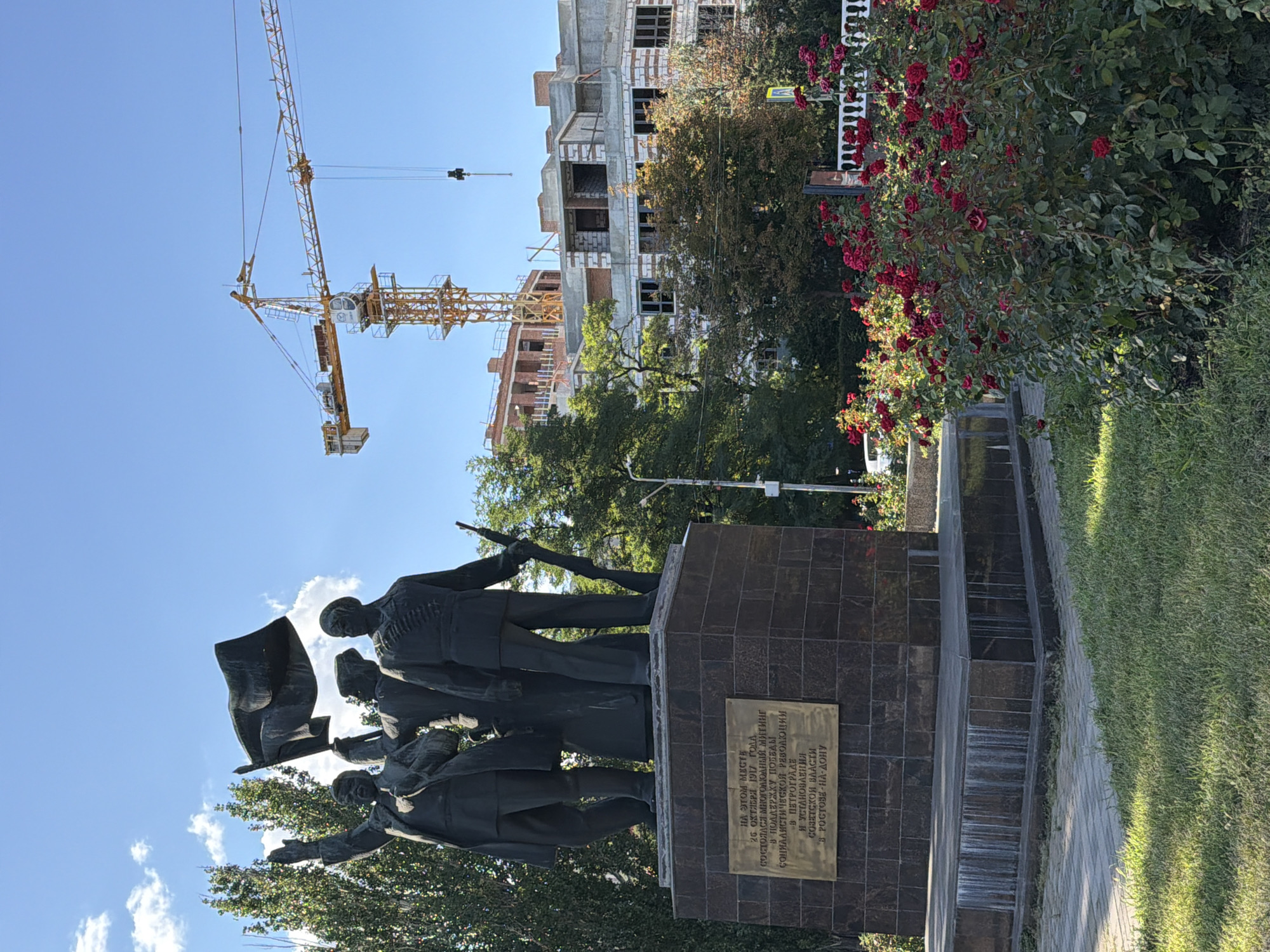 This is a medium-angle shot of a monument in Rostov-on-Don, Russia. The monument, a bronze statue of three men, stands on a dark stone pedestal. One man on the left raises his hand, holding a flag. The man in the center holds a rifle, and the man on the right is also holding a rifle. In the background, a yellow construction crane and a building under construction are visible against a clear blue sky. A bush of red roses is in the foreground on the right. The monument has a plaque on its base with Russian text. The area around the monument is paved with gray bricks and has a grassy area.
