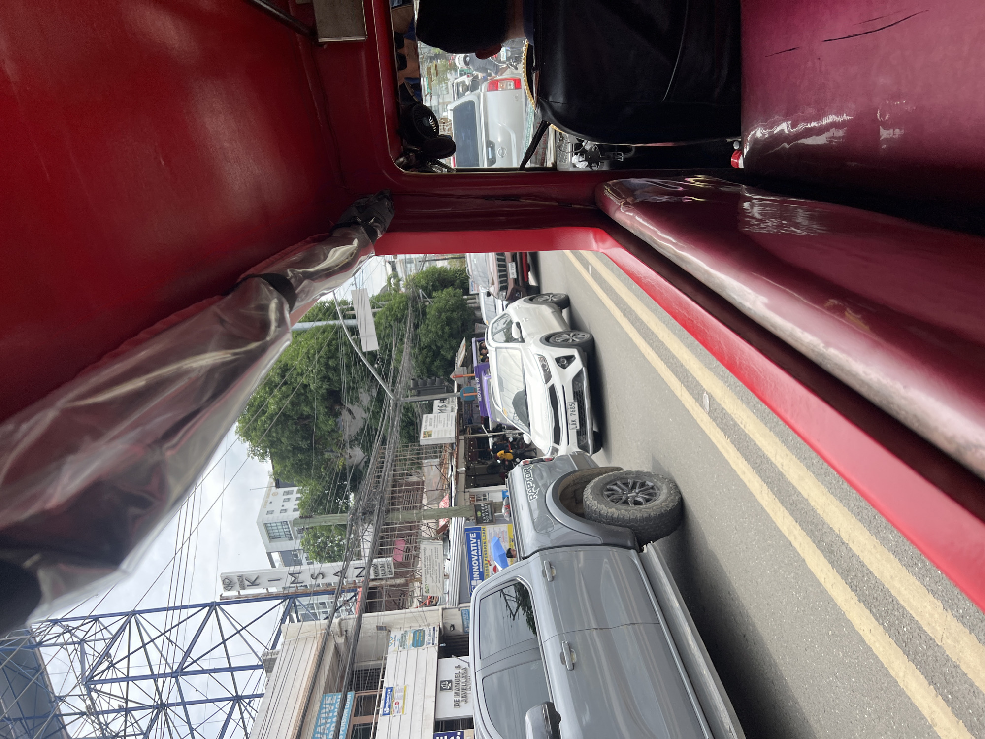 The image captures a street scene in Davao City, Philippines, viewed from inside a red vehicle. In the foreground, the red interior of the vehicle, possibly a jeepney or a similar public transport, is visible. The camera is looking out the side window onto a busy street.
On the left side of the street, a silver Ford Ranger Raptor pickup truck is prominently featured, with its large tires and "RAPTOR" branding visible. Beside it, a white Kia hatchback with license plate "LAK 7685" is also in traffic. Further down the street, other vehicles, including a silver SUV and a motorcycle, are present.
The background reveals buildings with various signage, indicating commercial establishments. One sign reads "AGENCIA KIMSAN," suggesting a financial institution or similar business. Power lines and traffic signals are visible, typical of an urban environment. Green trees can be seen interspersed between the buildings. The sky is overcast, suggesting a cloudy day. The road itself is asphalt with yellow lane markings. The overall impression is one of moderate urban traffic in daylight.