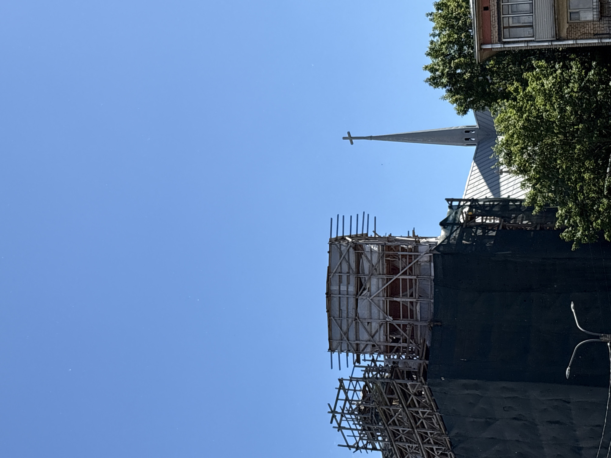The image shows a construction site in Saint Petersburg, Russia, on a clear, sunny day. A large building, possibly undergoing renovation, is partially covered by dark netting and scaffolding. The scaffolding is made of wood and metal, and it surrounds a section of the building that is also covered in white sheeting. In the background, a church steeple with a cross on top is visible, along with trees and the upper floors of another building. The time of day appears to be afternoon, given the angle of the shadows and the brightness of the sky. There is no text visible in the image. The overall scene suggests ongoing construction work in an urban environment.
