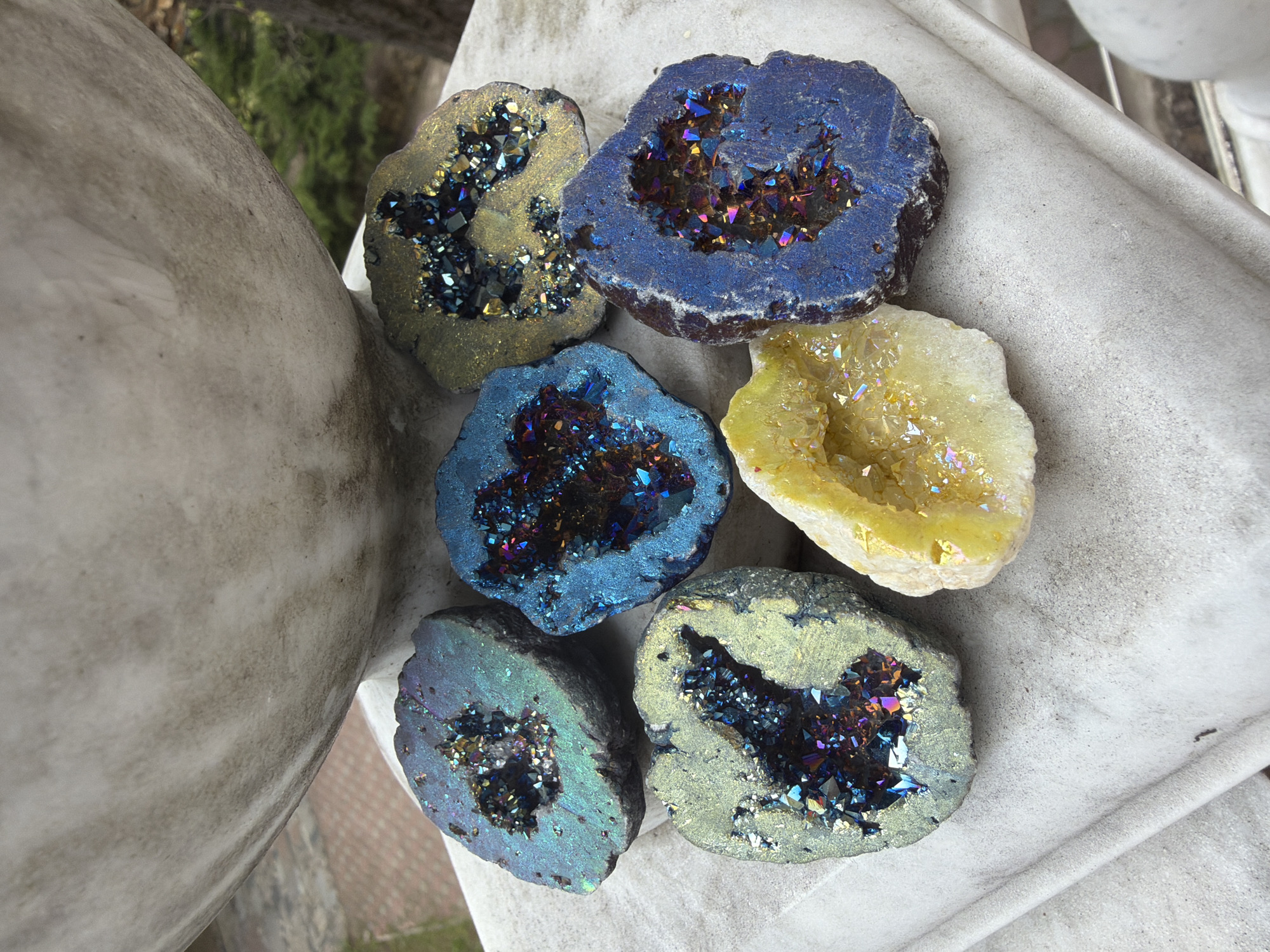 The image shows six halved geodes arranged on a white surface, likely a stone or concrete structure. The geodes are of various colors including gold, blue, and yellow, with iridescent crystal formations inside. The composition is set outdoors, as evidenced by the natural lighting and partial view of foliage and a sculpted stone object, potentially a statue or architectural element, in the background. The location is Unknown, Unknown.