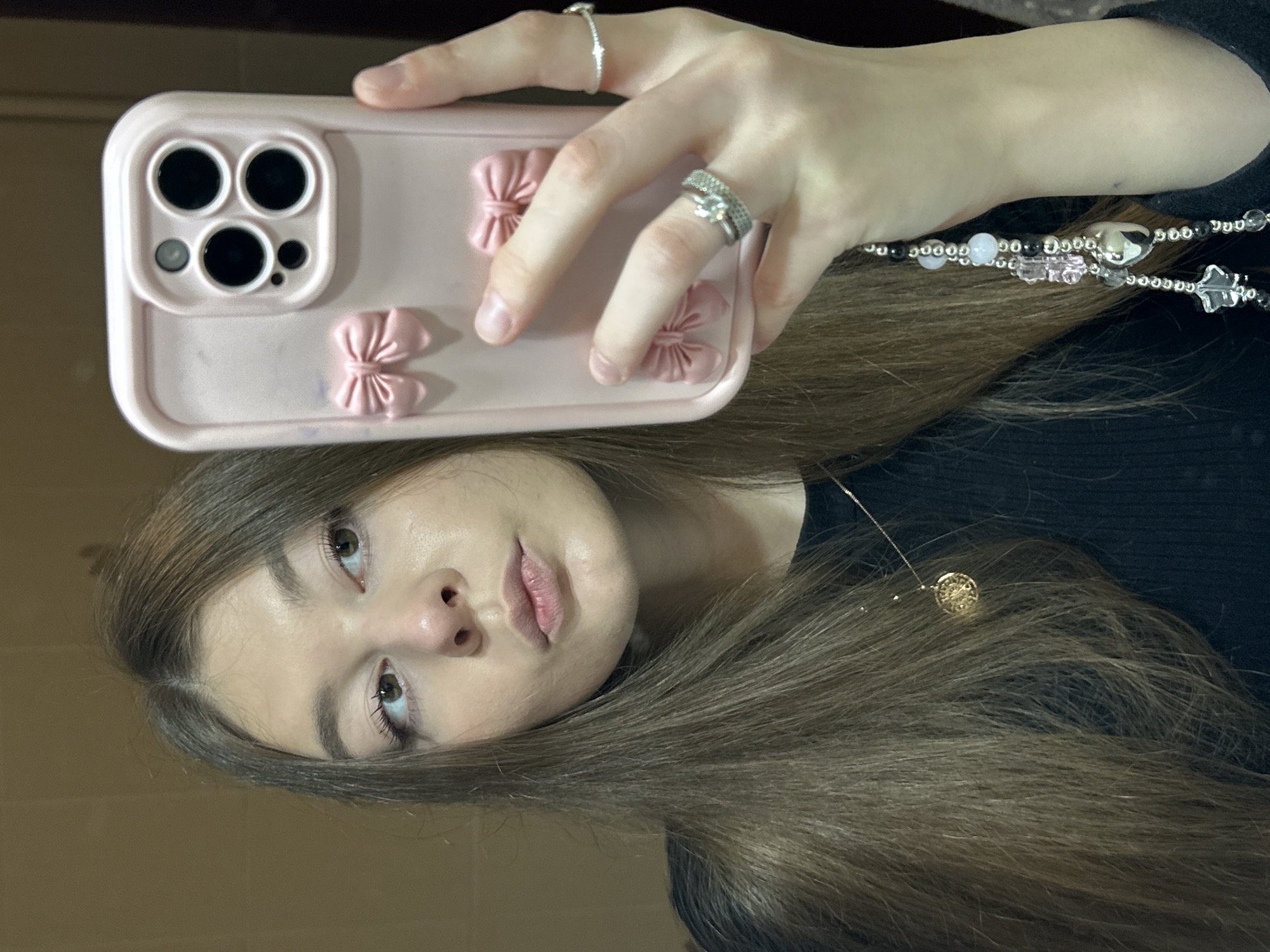 A young woman is captured taking a mirror selfie in an indoor setting, provided with the context of Kazan, Russia. Her head and upper torso are reflected in the mirror, with her long, dark brown hair flowing down her shoulders. She looks towards the camera with a neutral, slightly contemplative expression.
Her hand, adorned with a silver ring on her index finger and two stacked rings on her ring finger, holds a light pink smartphone. The phone's case is a prominent feature, decorated with several small, three-dimensional pink bows. The triple-camera system, including two larger lenses and one smaller lens, along with a flash, is clearly visible on the top left corner of the phone's back.
Around her neck, a delicate gold chain necklace with a small, round, intricately patterned pendant is visible. Additionally, a beaded necklace or phone strap, comprising silver, clear, black, and light pink beads, alongside a star-shaped and a heart-shaped charm, drapes from her hand or wrist over her dark clothing. The background visible in the reflection is a plain, warm, solid-colored surface, possibly a wall. There are no specific time of day cues or visible text within the image.