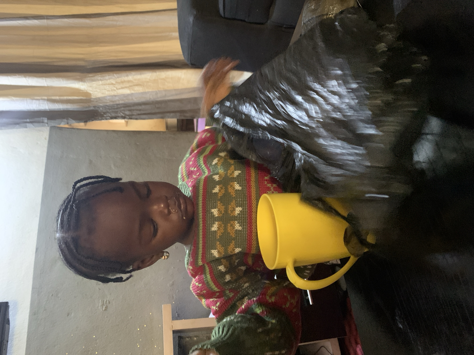 A young Black child with dark skin and braided hair is seated at a dark table, looking down at a black plastic bag. The child is wearing a colorful sweater with a knit pattern of green, red, and white. A bright yellow mug with a handle is positioned to the child's left. The background includes a light-colored wall, a tan curtain, and a dark sofa. The child's face is partially obscured by shadows, but a bit of food residue is visible around their mouth. The scene appears to be indoors in what could be a residential setting.
