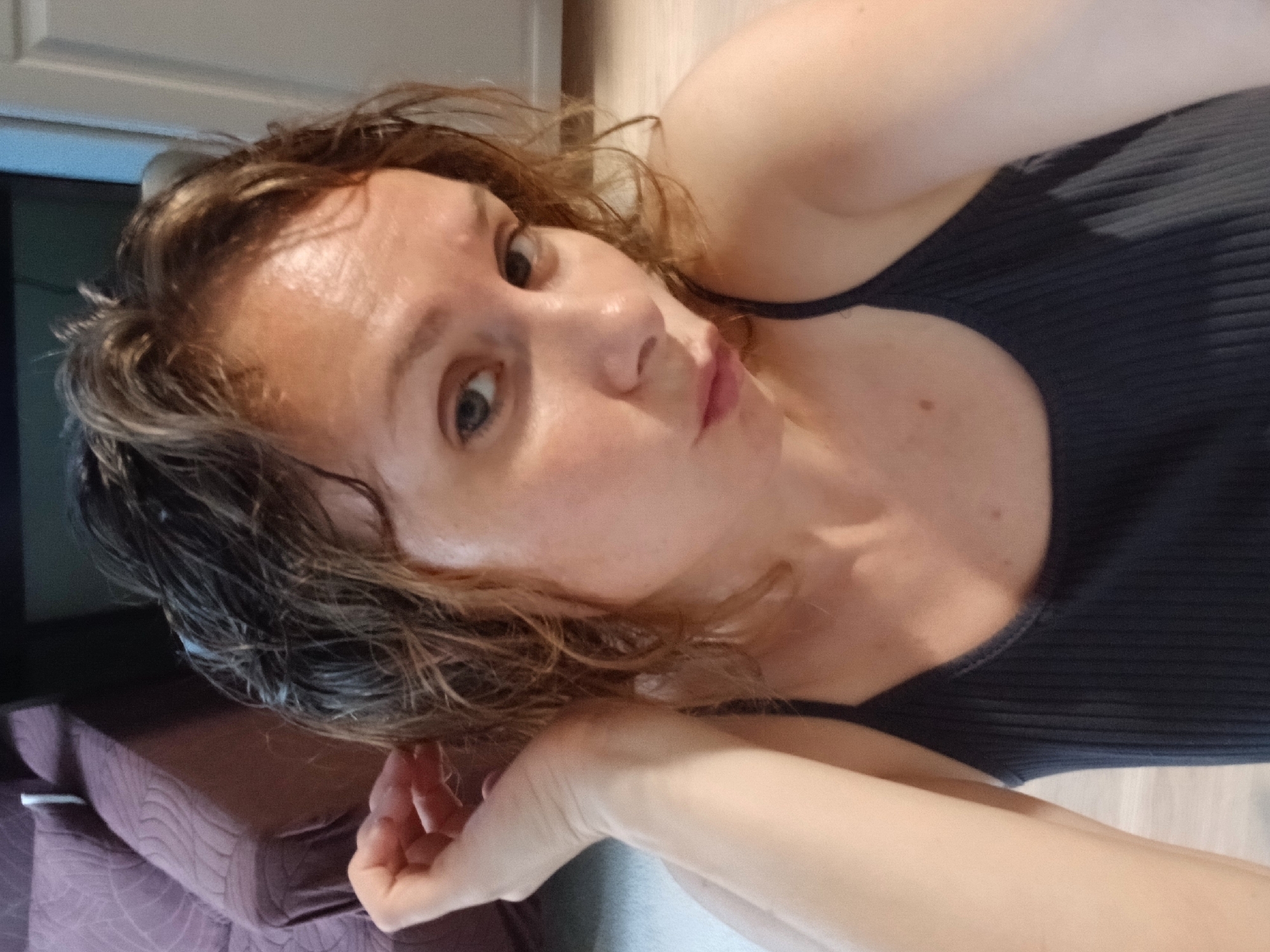 A close-up, waist-up selfie of a Caucasian female with wet, medium-length brown hair styled loosely. She has blue eyes, fair skin, and is wearing a dark gray ribbed tank top. Her left arm is bent, with her left hand resting near her head, touching her hair. The background is blurred but appears to be an indoor domestic setting. To the left, a portion of a purple sofa with a textured pattern is visible. To the right, a white vertical surface, possibly a door or wall, and a darker vertical surface, possibly a cabinet, are present. The lighting suggests artificial indoor light. The image was taken in Yekaterinburg, Russia.