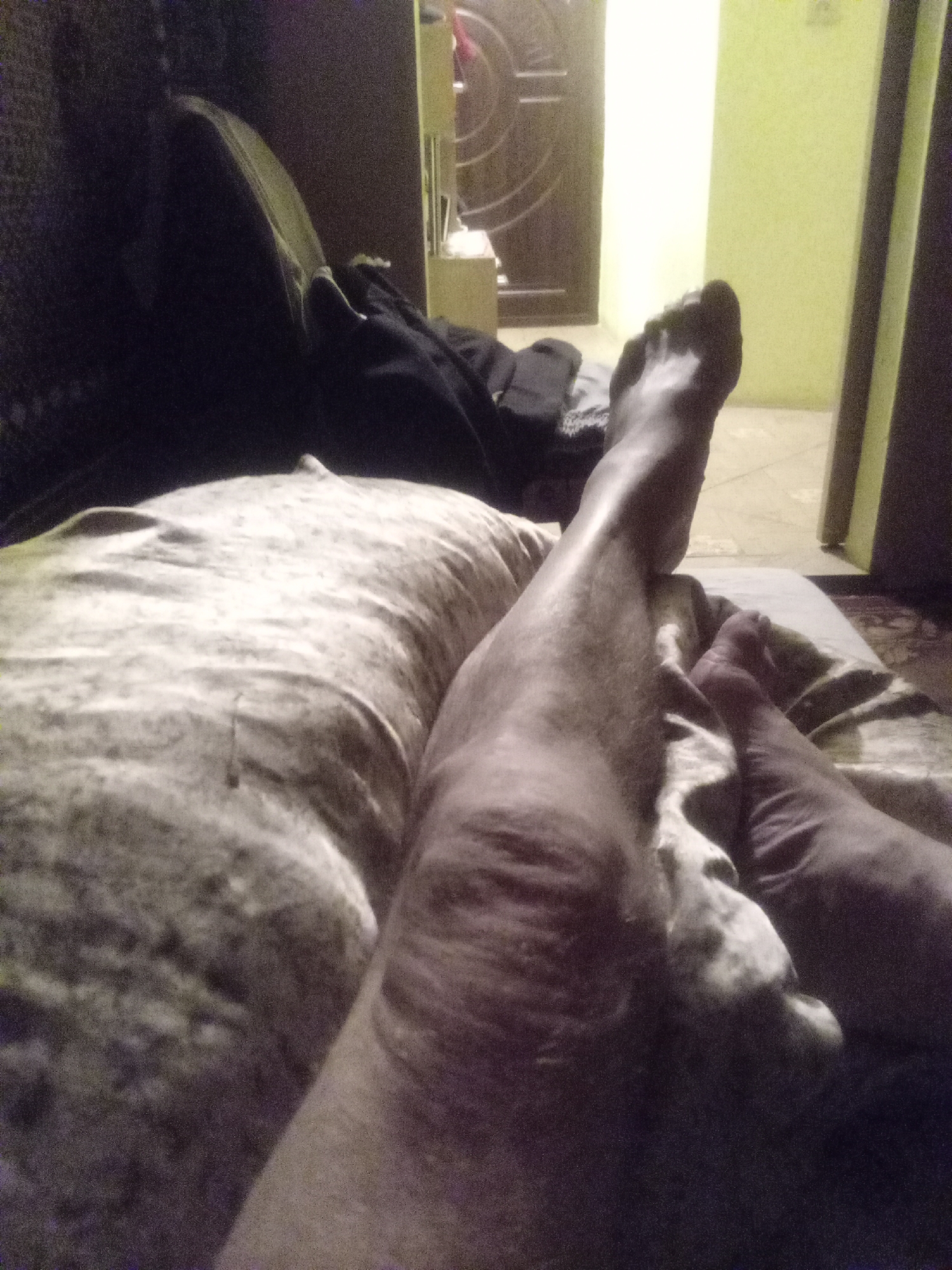 The image shows a person lying on a bed or couch, with their legs and feet prominently in the foreground. The person appears to be male, based on the visible skin and hair on the legs. Their left leg is bent at the knee, and the right leg is extended. The feet are bare. The background is dimly lit, with what appears to be a door and a wall painted in a light green color. There is also a dark, possibly upholstered surface to the left of the frame, which might be the back of the couch or a chair. Clothing, dark in color, is visible behind the person's legs. The overall impression is of a casual, indoor setting. No specific identifiable landmarks or features are visible to indicate the location within Tiachiv, Ukraine.