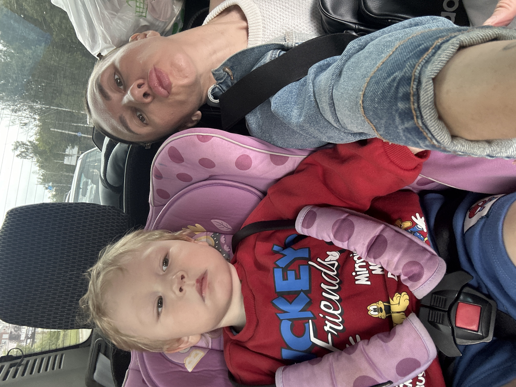 The image, taken inside a vehicle, features an adult and a young child, likely traveling. The perspective is from above, looking down on both subjects, who appear to be lying back or reclining in their seats.
The adult, positioned in the upper portion of the frame (when oriented correctly), has light skin and short, dark hair. They are wearing a light-colored, textured sweater, a denim jacket, and a black seatbelt across their chest. Their lips are pursed, and they look directly at the camera. A white plastic bag, possibly from a grocery store, is visible in the background above their head. A black bag with "ADIDAS" written on it is also visible behind the adult's shoulder.
Below the adult, a young child with light blonde hair and fair skin is seated in a purple car seat with pink polka-dotted shoulder strap covers. The child wears a red long-sleeved shirt with the words "MICKEY friends" prominently displayed in blue and white lettering, along with what appear to be Disney characters like Pluto and Minnie Mouse. The child's expression is neutral as they look towards the camera. The black buckle of their car seat harness is visible over their shirt.
Through the vehicle's rear window, trees and what seem to be other vehicles or buildings can be seen, indicating an outdoor setting, likely daytime. The scene suggests a shared moment, possibly a selfie taken during a car journey in Izhevsk, Russia.