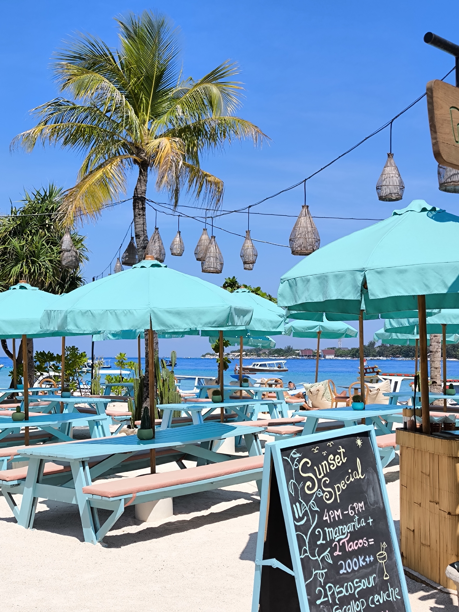 The image shows a beachside scene, likely at a resort or restaurant, with a tropical ambiance. The location is Unknown, Indonesia.
In the foreground, there are several light blue picnic tables with salmon-colored bench cushions, arranged on white sand. Teal-colored umbrellas provide shade over the seating area. A black chalkboard sign with a teal frame advertises a "Sunset Special" from 4 PM to 6 PM, including a deal on margaritas, tacos, and a combination of Pisco Sour and Scallop Ceviche.
In the background, a clear blue ocean is visible, dotted with boats. A few people can be seen relaxing in chairs. A tall palm tree stands to the left, and decorative wicker lanterns are suspended from strings overhead. The sky is clear and bright blue.