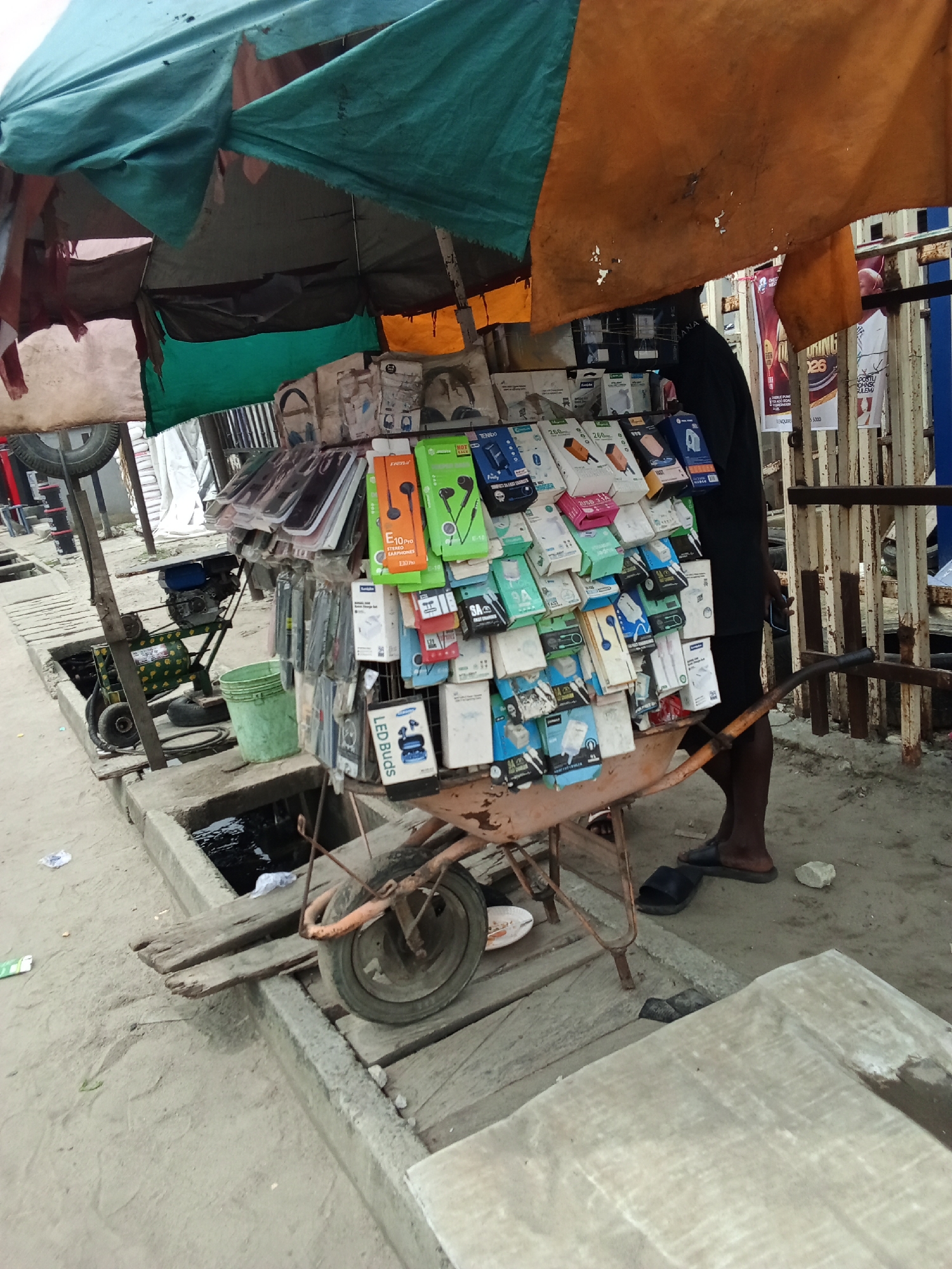 The image depicts a mobile vendor in Ado, Nigeria, selling a variety of phone accessories displayed on a rusty wheelbarrow. The accessories include headphones, earbuds, phone cases, and chargers. The vendor, an African man in a black shirt, stands near the wheelbarrow. A makeshift canopy of green and orange fabrics provides shade. The scene takes place on a paved street with a drainage system. A generator is nearby, and posters hang on a metal fence in the background.