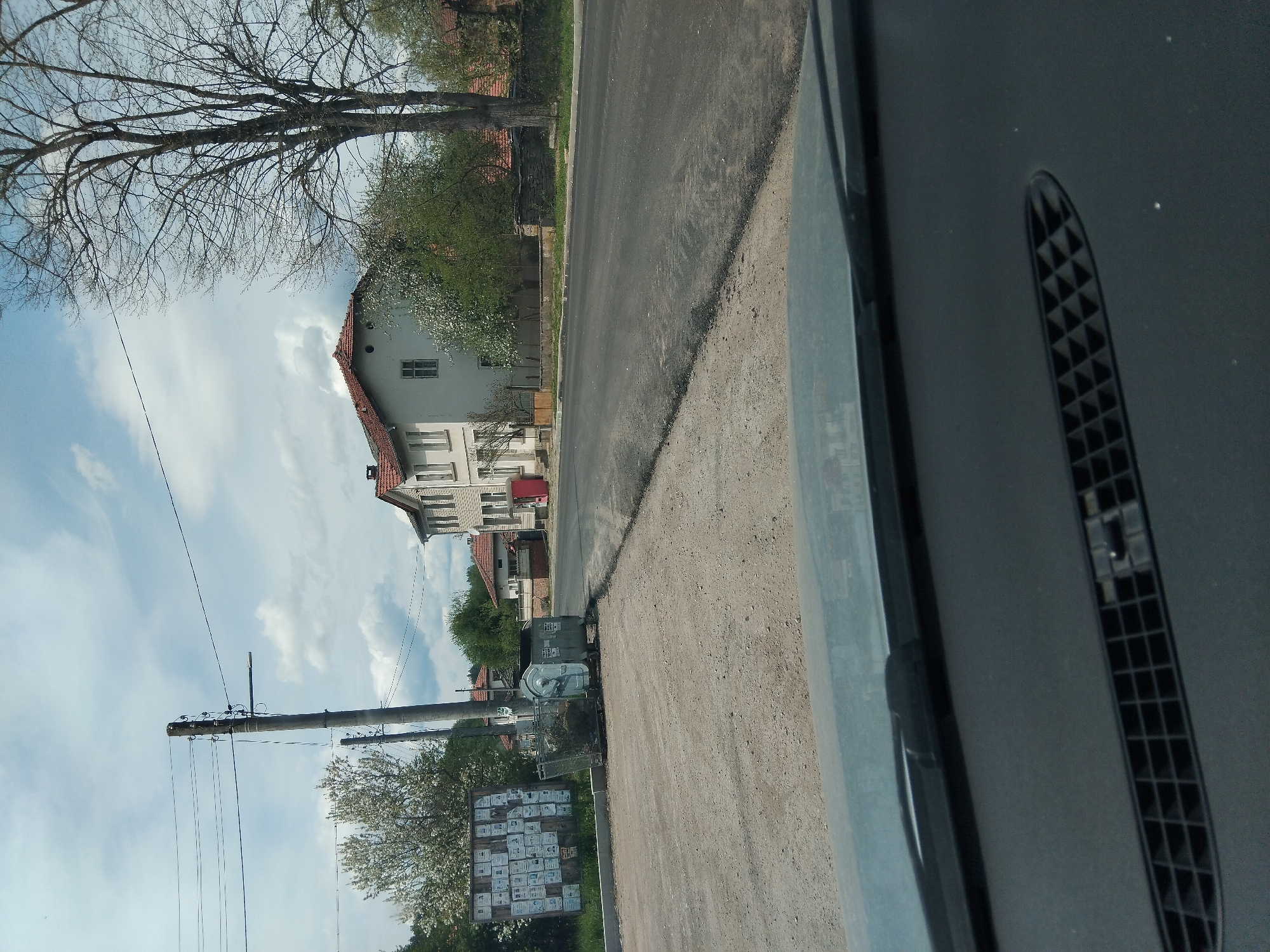 View from inside a vehicle looking out onto a street in Velchevo, Bulgaria. The street is paved, with a dirt shoulder. On the left side of the street, there is a building with a red roof. Trees with bare branches are visible. Power lines and a utility pole are also visible. A large sign with what appears to be many individual notices is present. A metal dumpster is next to the sign. Part of the vehicle's dashboard and vent are visible on the right side of the frame. The sky is blue with some clouds.