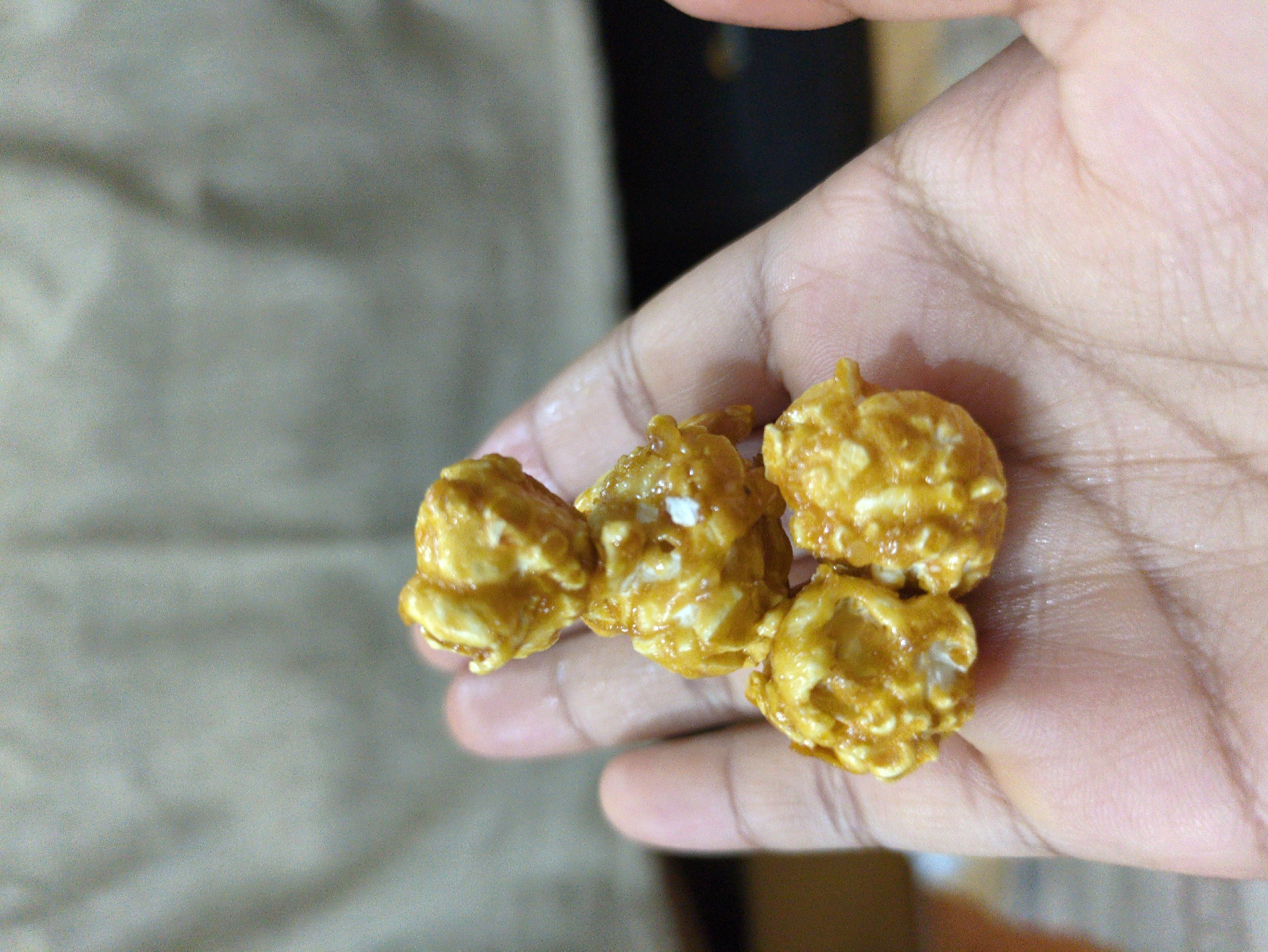 A close-up, eye-level shot captures a person's hand holding a small stack of caramel popcorn. The popcorn pieces are clustered together, with one piece resting on top of two others, which in turn are resting on a larger cluster. The caramel coating gives the popcorn a shiny, golden-brown appearance, with some visible white flecks of what appears to be popped corn. The hand, which is dark-skinned, is open with the palm facing upwards, providing a backdrop for the popcorn. The background is blurred, showing a textured, light-colored fabric, possibly a curtain or backdrop, and a darker, out-of-focus area that could be a wall or furniture. The image does not provide any specific visual cues to indicate a location in Bhagsu, India, or any other city.