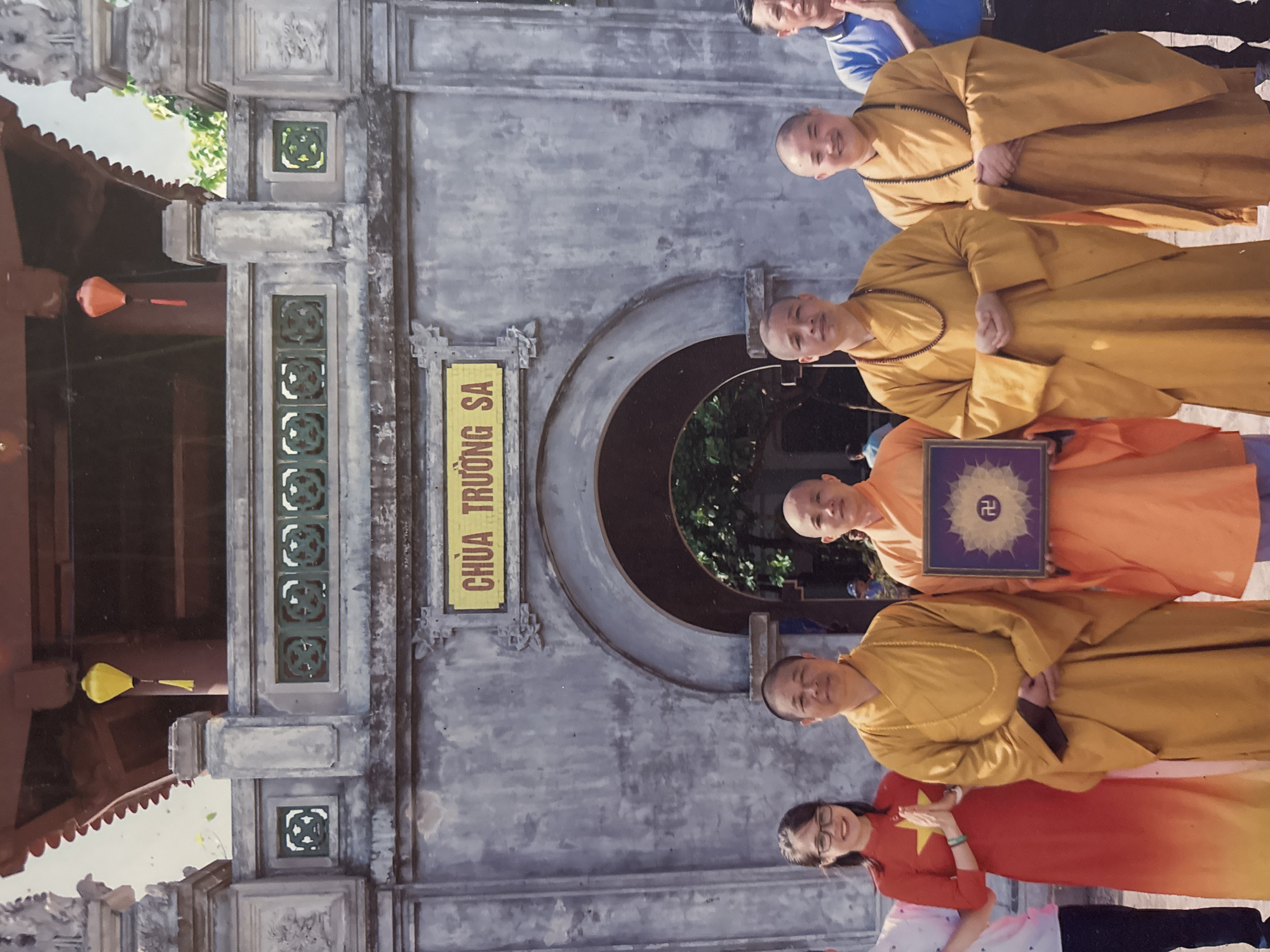 A group of six individuals, five in Buddhist monk robes of yellow and orange, and one woman in a red and yellow dress, stand in front of a weathered, ornate stone gateway with a sign above it that reads "CHÙA TRƯỜNG SA". Two yellow and one red lantern hang from the eaves of the structure. The individuals are posing for a photograph. The woman on the far left is wearing glasses and has her hands clasped in a prayer gesture. She is wearing a red dress with a yellow star on the chest, and it fades to yellow at the bottom. The monks are all bald and wear necklaces of beads. One monk, dressed in orange, is holding a square, framed object with a swastika symbol in the center. The gateway appears to be the entrance to a temple or religious site located in Tây Hoa Lư Ward, Vietnam. The overall scene is bright and appears to have been taken outdoors on a clear day.