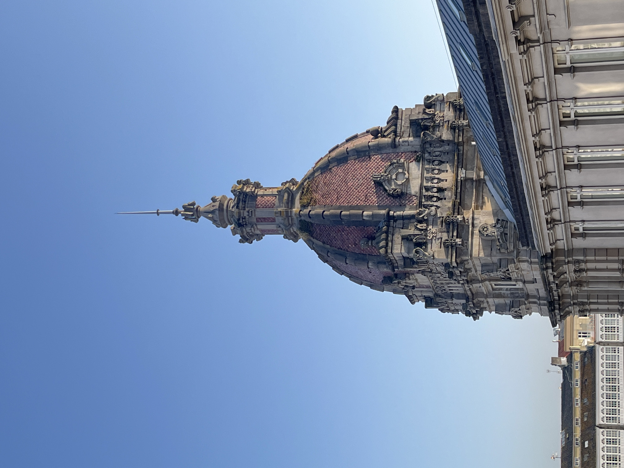 The image captures a low-angle, daytime view of the dome of the Banco Pastor building in A Coruña, Spain, juxtaposed with a modern glass and steel structure. The dome features reddish-brown tiling, ornate stone carvings, and a spire. Adjacent to the dome, a modern building exhibits a linear pattern of glass panels and metallic trim. The sky is clear and blue, and the angle of the shot includes glimpses of other buildings in the background.