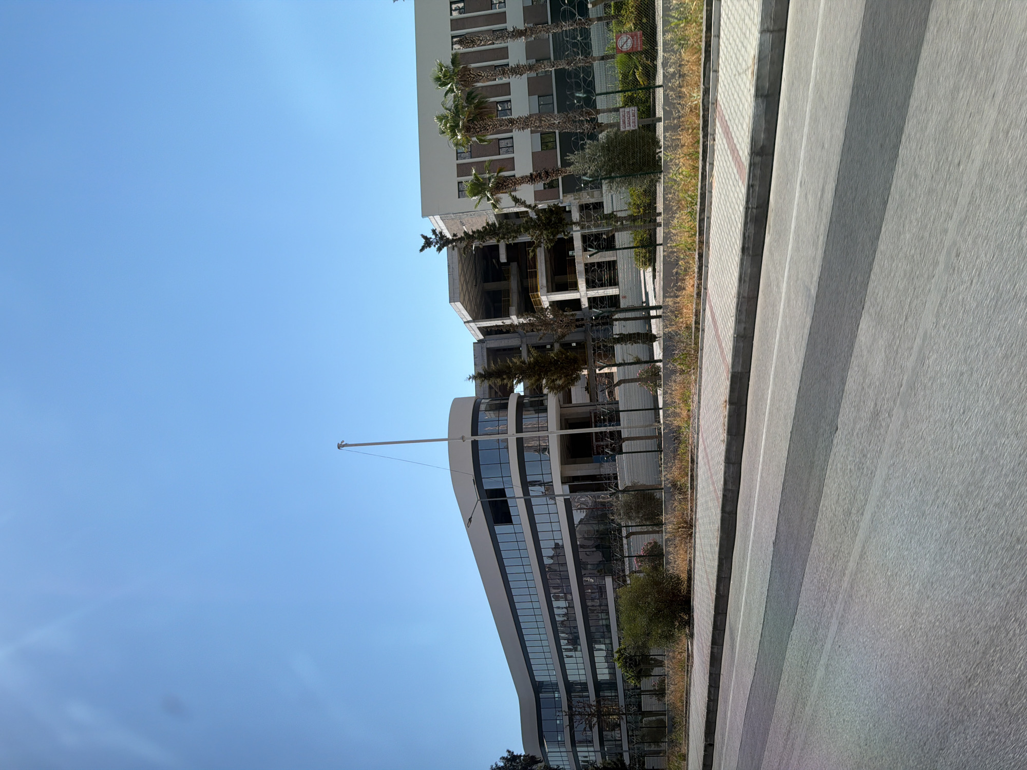 The image shows a modern building under construction next to an existing building. The scene is outdoors, with a clear blue sky suggesting a sunny day. The foreground is dominated by a gray paved surface, possibly a sidewalk or road, with diagonal stripes that create a textured appearance. To the left of the paved area, there's a strip of dry grass and then a fence.
Beyond the fence, the buildings are the main subjects. The building on the right appears to be complete, with multiple floors featuring rows of windows and what looks like palm trees in front of it. The building on the left is clearly under construction, with its internal framework exposed. Several trees, including some with bare branches, are visible around this structure.
A tall flagpole stands prominently in front of the unfinished building, reaching high into the sky. The buildings are modern in design, with the completed section featuring a curved facade with large glass windows.
There is some visible text on a sign attached to the fence in the middle-right of the image. It is difficult to read clearly due to the resolution and angle, but it appears to be a warning sign, possibly related to construction or safety. A red circular symbol with a line through it is also visible on the sign, often used to indicate prohibition.
The overall impression is one of urban development in a sunny climate, with a contrast between a finished modern structure and a new one in progress. The scene is static, with no people visible. The time of day is likely midday or afternoon due to the bright sunlight and clear sky. The location context of Konyaaltı, Turkey, suggests a Mediterranean or coastal environment, consistent with the presence of palm trees.