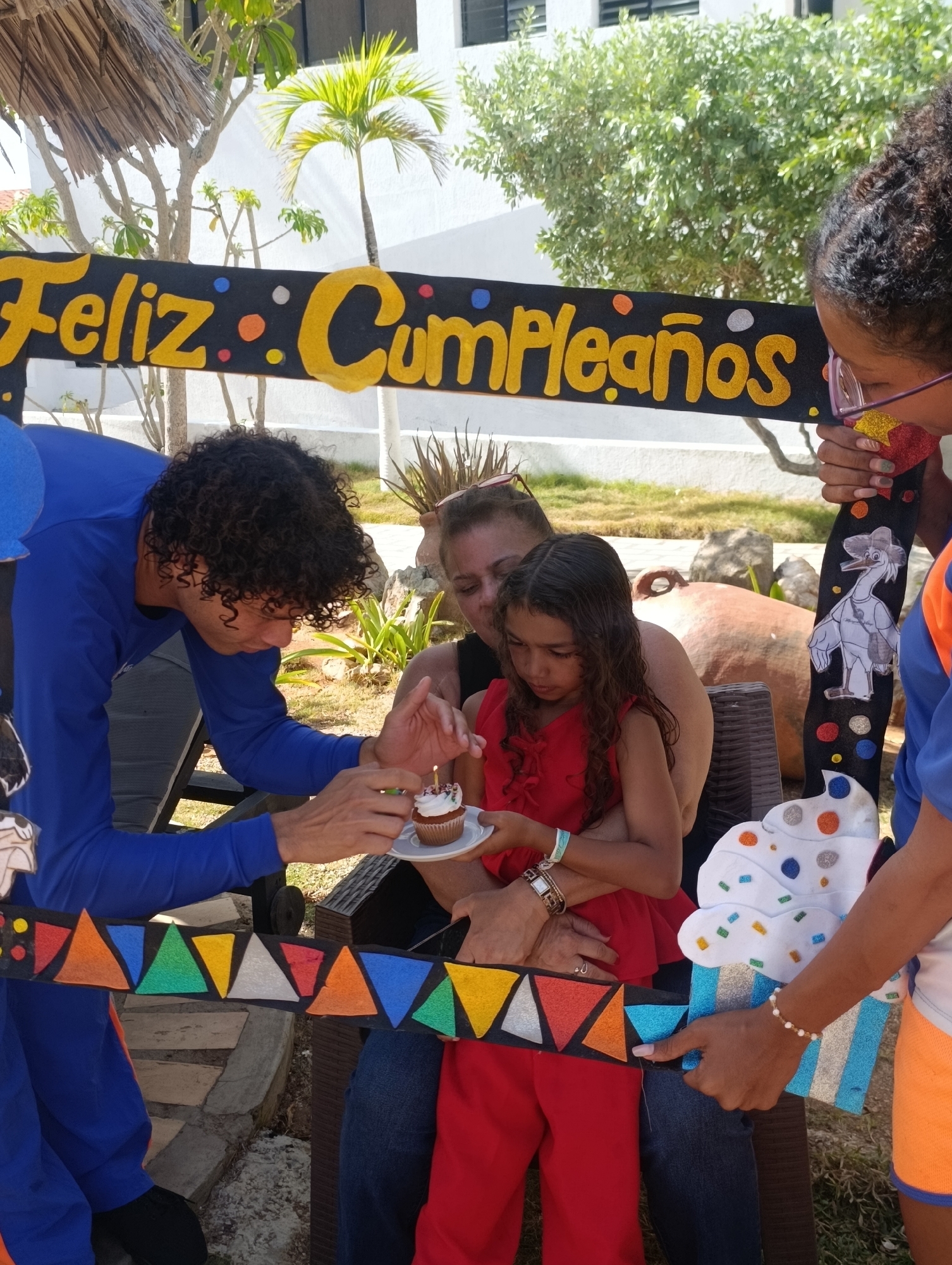 The image captures a birthday celebration, with a decorative banner proclaiming "Feliz Cumpleaños" (Happy Birthday in Spanish) in yellow lettering on a black background.
A young girl in a red outfit sits on a woman's lap, likely her mother. A person in a blue long-sleeved shirt holds a cupcake with a lit candle. Another person in a blue shirt and orange shorts holds the right side of the "Feliz Cumpleaños" banner.
The celebration appears to be outdoors. The scene is in Porlamar, Venezuela, identifiable by the language of the banner.