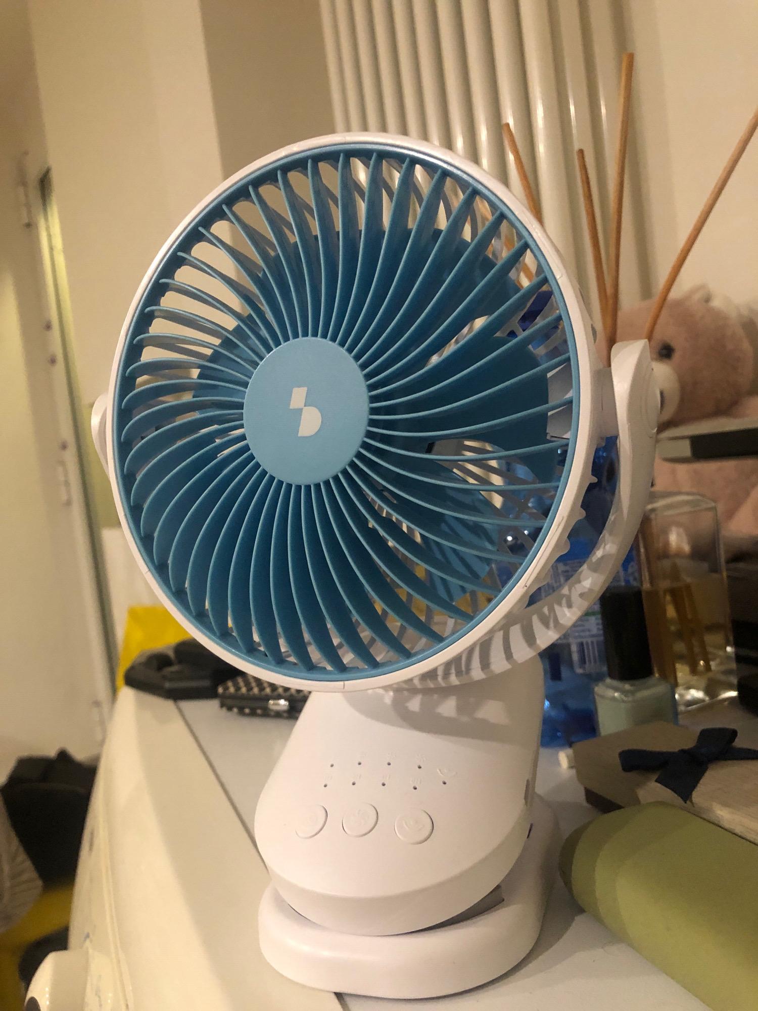 A close-up, eye-level, indoor shot shows a white and blue portable clip-on fan positioned on a white surface, likely a dresser or table. The fan's blades are a vibrant blue, and its base and clip are white. The fan is facing slightly to the left. In the background, a blurred pink teddy bear sits behind the fan. To the right of the fan, two small glass bottles are visible, one containing a dark liquid and the other a lighter, amber-colored liquid. A gift-wrapped box with a navy blue ribbon lies in front of the bottles. Behind the fan, vertical blinds are visible. The lighting suggests an indoor environment, possibly a bedroom or living area. The scene is devoid of any indications of the city of San Pietro in Casale, Italy.