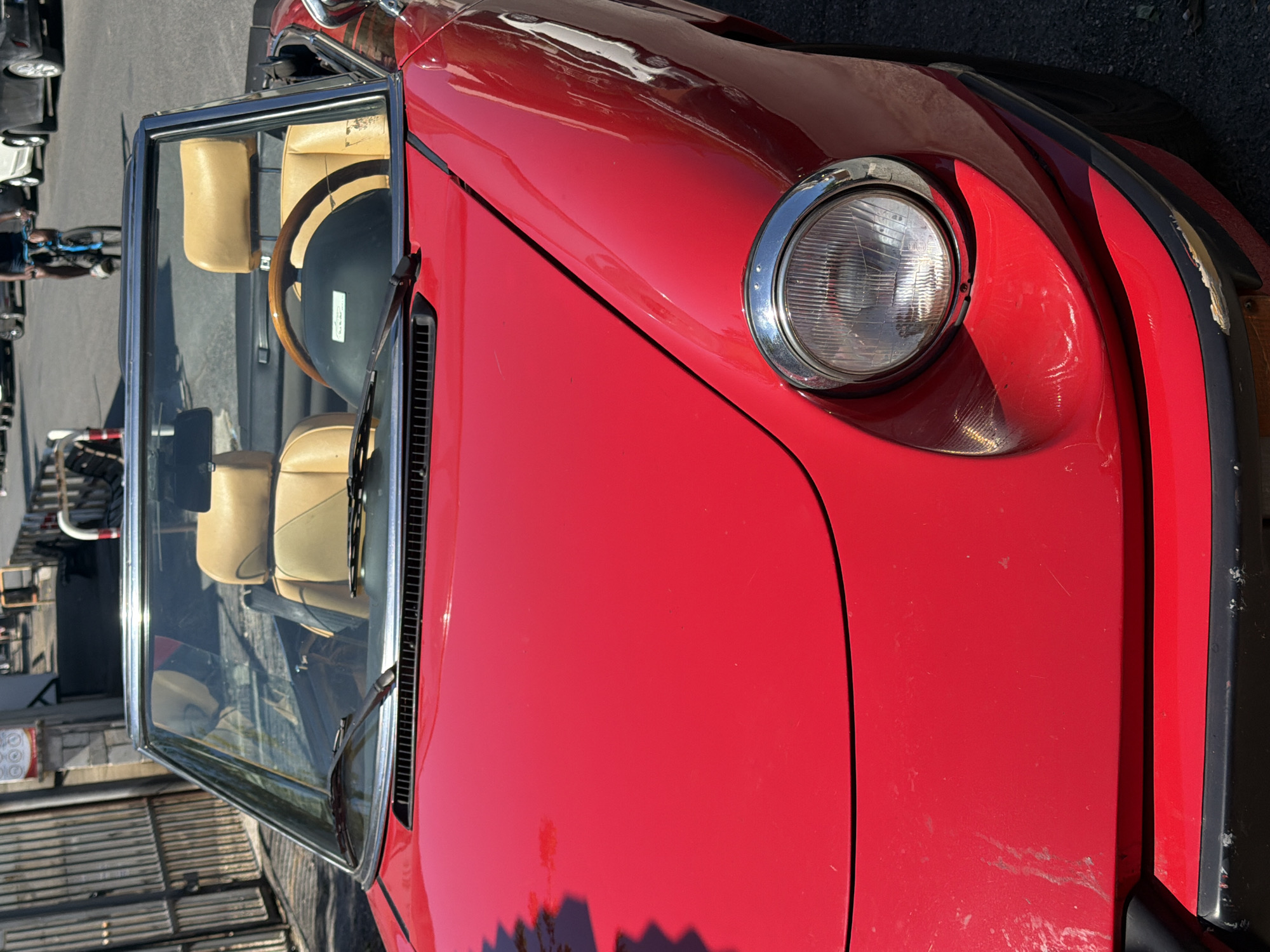 An exterior, eye-level, medium shot captures the front of a bright red, vintage convertible car parked on a paved street in Rome, Italy. The car's headlights, hood, and windshield are visible. The interior features light tan leather seats and a wooden steering wheel. In the background, a person is riding a bicycle, and other vehicles and buildings are visible. The overall scene is illuminated by bright daylight.