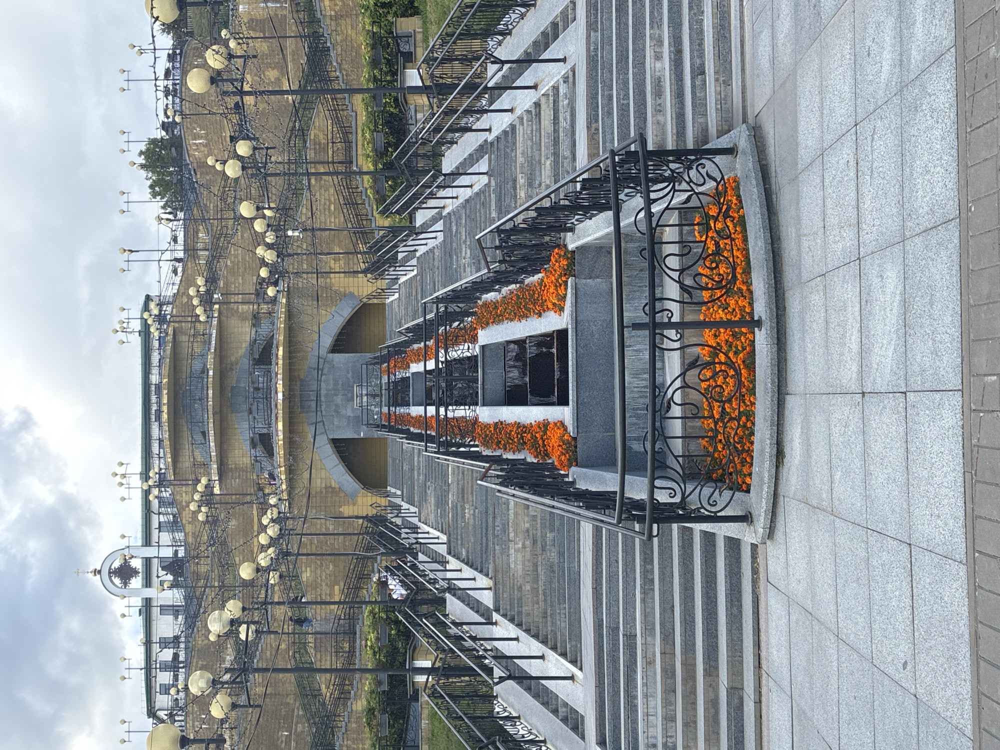 The image shows a wide stone staircase adorned with numerous black wrought-iron railings and posts. Along the sides of the staircase, rows of vibrant orange flowers are planted, creating a colorful border. At the base of the staircase, a paved area with large, light grey square tiles is visible. In the background, a tiered structure with stone walls and multiple levels of stairs is topped by a white building with a green roof and a distinctive arched structure with a cross, suggestive of a religious institution, possibly the Church of the Transfiguration in Mahilyow, Belarus. Several streetlights with yellow globe-shaped lamps are mounted on decorative black poles, lining the upper tiers of the structure. The sky is overcast with grey clouds, and the overall atmosphere is somber. There are no discernible people actively engaged in any specific actions within the frame.