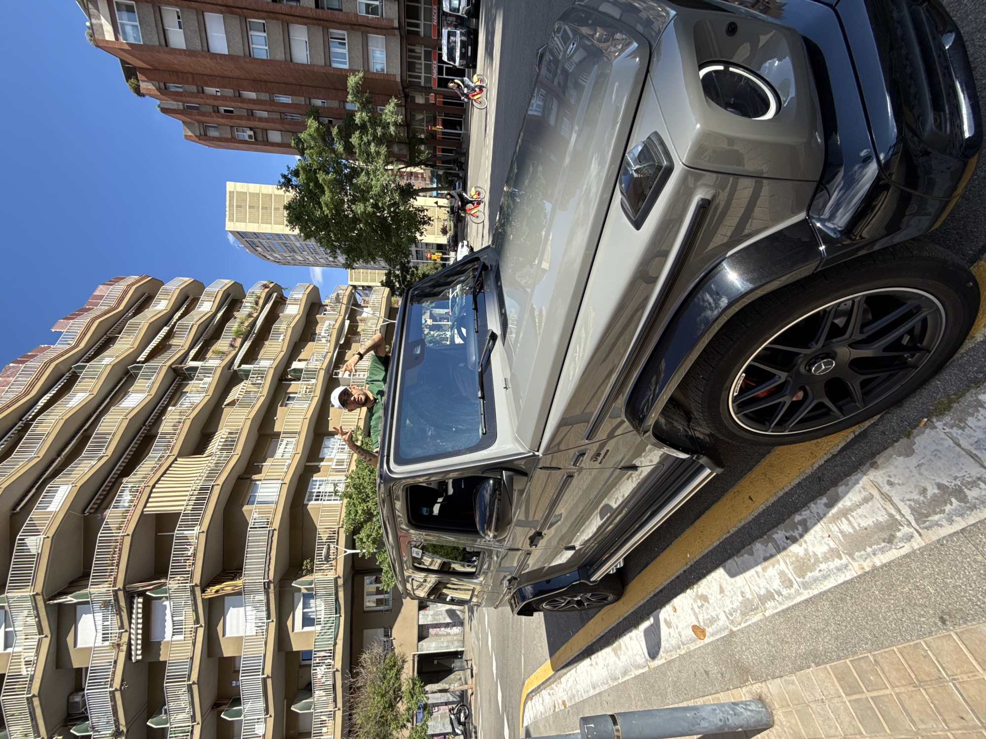 A gray Mercedes-Benz G-Class SUV is parked on a city street with a man leaning out of the driver's side window, making a hand gesture. The vehicle is partially on the sidewalk next to a yellow line. Buildings with a distinct architectural style, marked by curved balconies, are in the background. Other buildings of various designs are also visible, suggesting an urban setting. The scene is bathed in bright sunlight, casting strong shadows. A few cyclists and other parked vehicles can be seen further down the street. The location is likely Barcelona, Spain, based on the architectural style of the buildings.