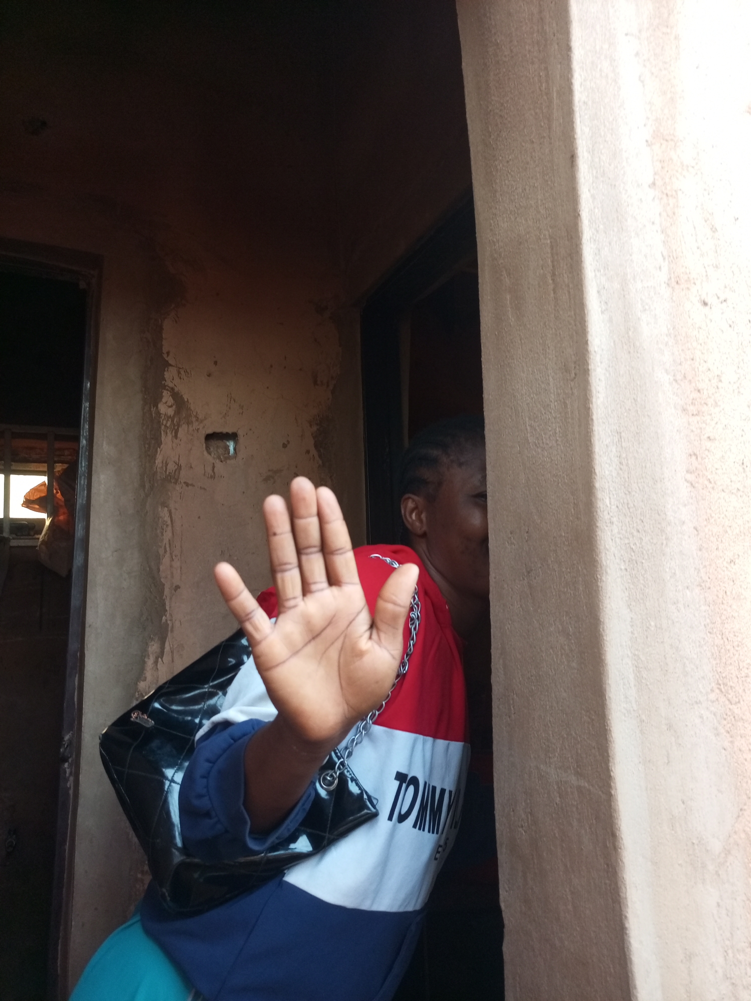 The image depicts a young Black woman standing in a doorway, holding up her left hand in a "stop" gesture towards the camera. She is wearing a red, white, and blue color-blocked sweatshirt with the "TOMMY" logo visible on the chest, and a shiny black quilted handbag with a silver chain strap is slung over her left shoulder. Her hair is braided and pulled back. The background shows the rough, textured walls of what appears to be a building with a dark interior visible to the left, and a lighter, possibly sunlit area beyond that. The right side of the frame is dominated by a light-colored, textured pillar or wall. The scene is indoors, likely within a residential or commercial structure. The location is identified as Tempure, Nigeria.