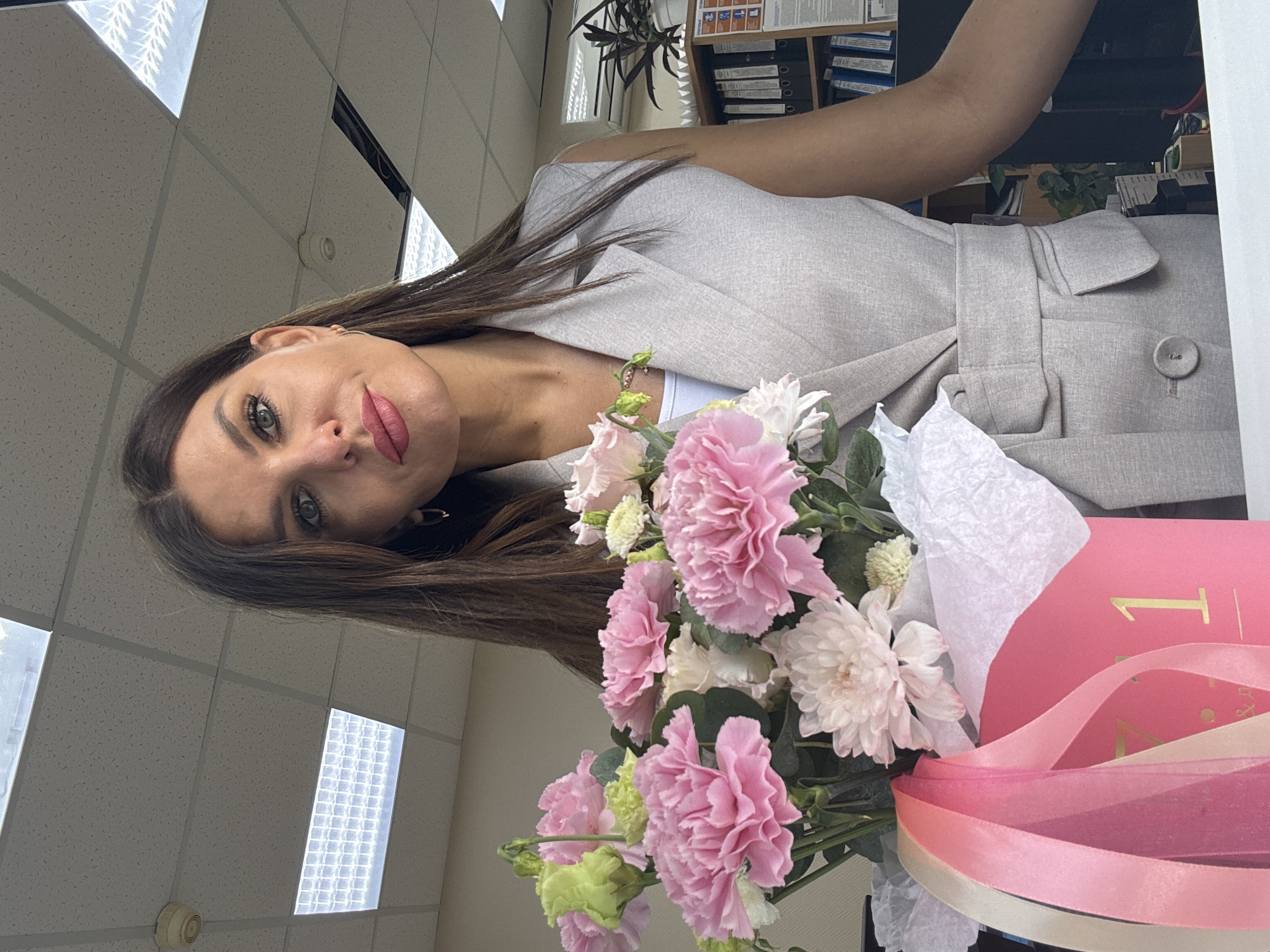 A woman is holding a bouquet of pink and white flowers, along with pink ribbons and a pink box with a partially visible "1" in gold. She is wearing a light-colored blazer. The background features a ceiling with fluorescent lights and a shelf with binders and plants. This photo may have been taken in Chaikovsky, Russia.