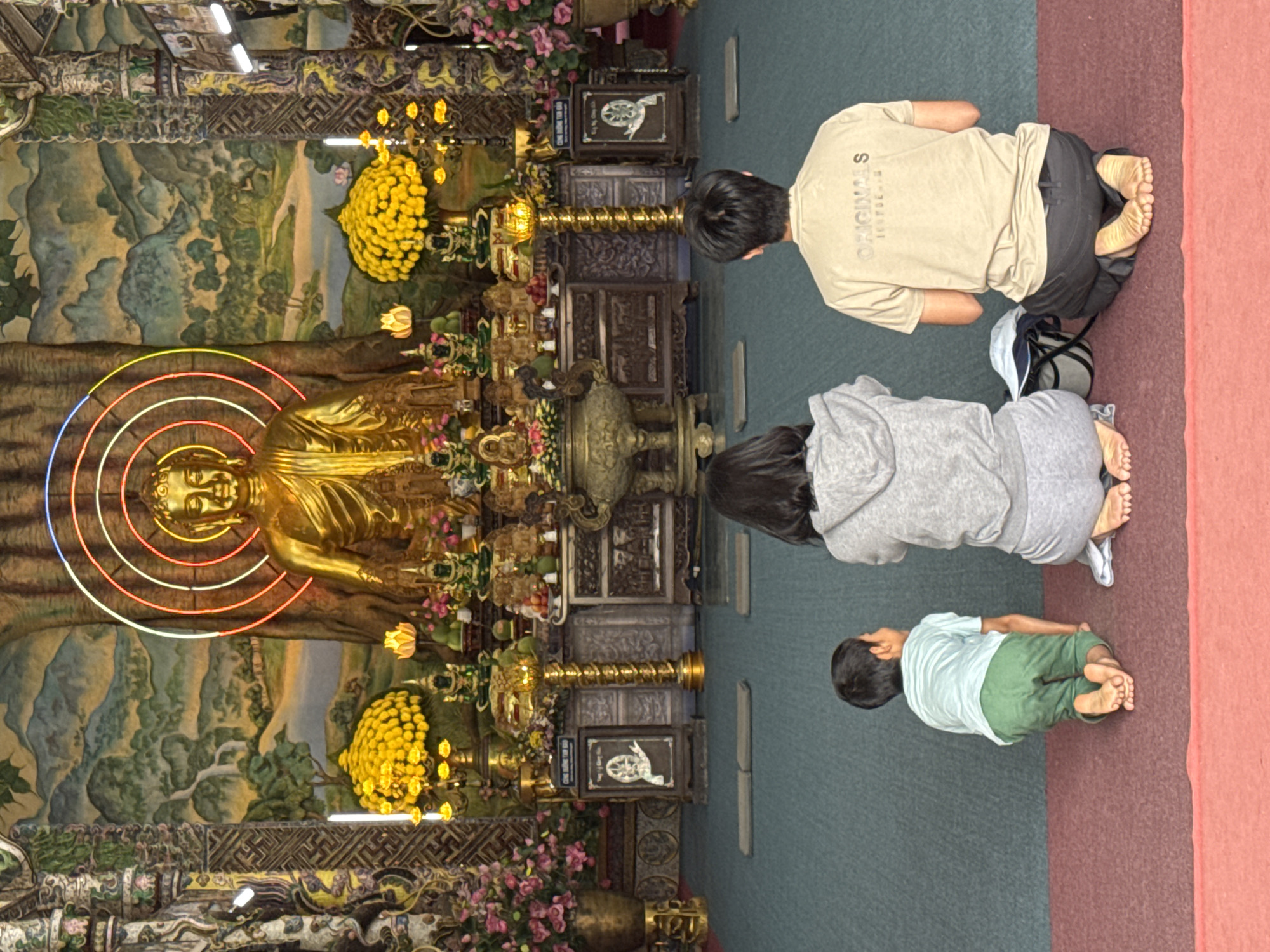 The image depicts a religious scene within a Buddhist temple in Xuân Thành, Vietnam. Three individuals are kneeling on the floor in prayer, facing a large golden Buddha statue. The Buddha is positioned centrally in the frame, adorned with a colorful neon halo. To the left and right of the Buddha are floral arrangements and smaller religious iconography. The temple interior features ornate decorations and a painted mural on the backdrop depicting a landscape. The floor is covered with a carpeted surface. The individuals are a young boy in a light blue shirt and green pants, a woman in a grey hooded sweatshirt and grey pants, and a man in a tan t-shirt and dark pants. Their posture indicates they are engaged in prayer or meditation.