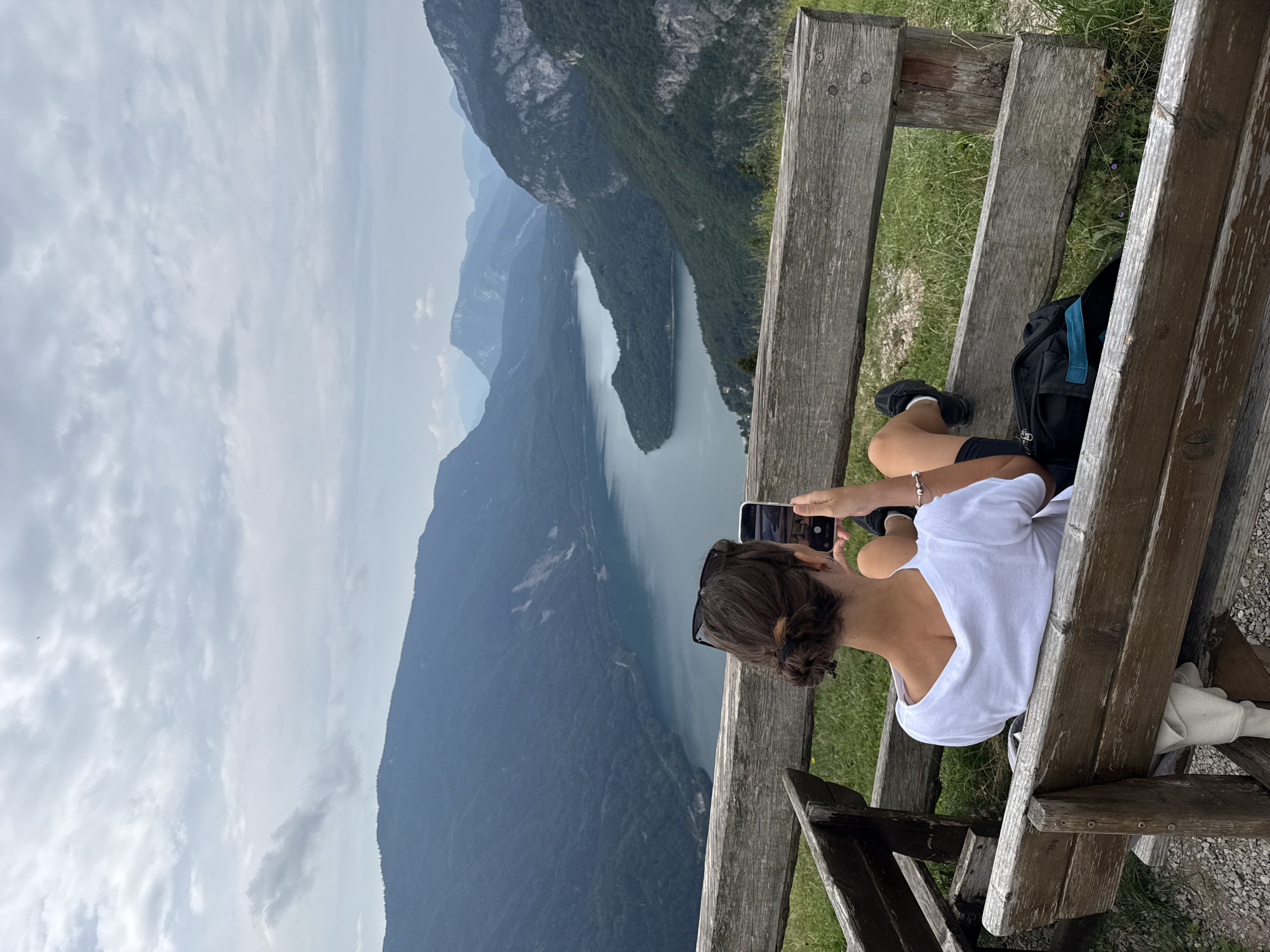 A person is sitting on a rustic wooden bench on a grassy overlook, looking at a smartphone. The person is seen from behind, wearing a white t-shirt and black shorts, with dark hair tied up in a bun. The bench faces a vast mountainous landscape with a large, serene lake nestled between the peaks. The sky is overcast with dramatic clouds. This viewpoint is characteristic of the panoramic vistas found around Molveno, Italy, offering expansive views of Lake Molveno and the surrounding Brenta Dolomites.