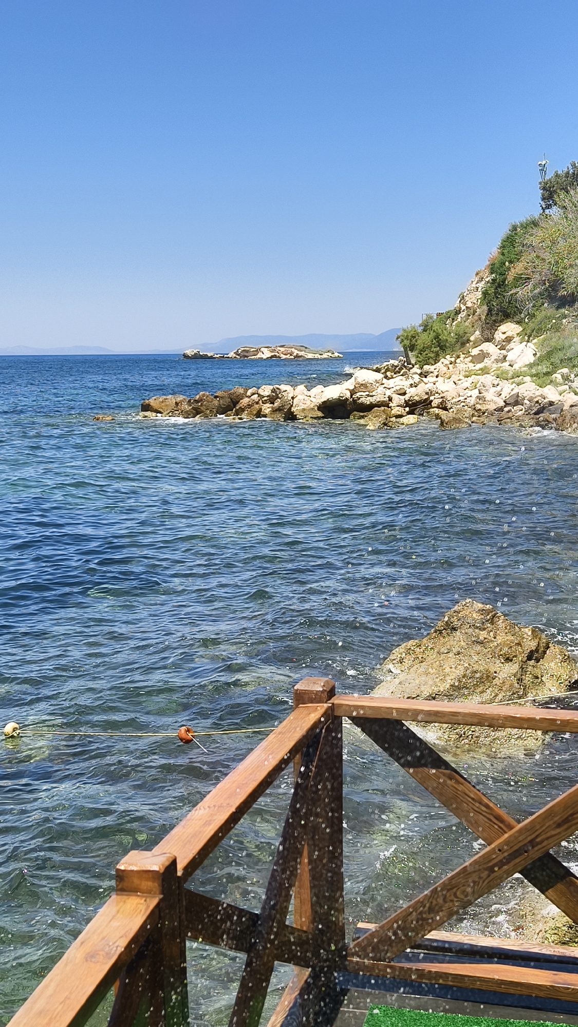 The image depicts a coastal scene in Kuşadası, Turkey, on a clear, sunny day. The foreground features a wooden railing, possibly part of a pier or boardwalk, with water lapping against the rocks below. The water is a vibrant blue, with visible ripples and some small whitecaps. In the middle ground, a rocky shoreline extends into the sea, with larger boulders and smaller stones. A few tufts of green vegetation cling to the rocks. In the distance, a small, uninhabited island with sparse vegetation rises from the water, and further beyond, the hazy silhouette of the mainland coastline is visible under the bright blue sky. A yellow buoy and a smaller orange marker are visible in the water, suggesting a possible mooring or boundary line. The overall impression is one of a serene and natural seaside environment.